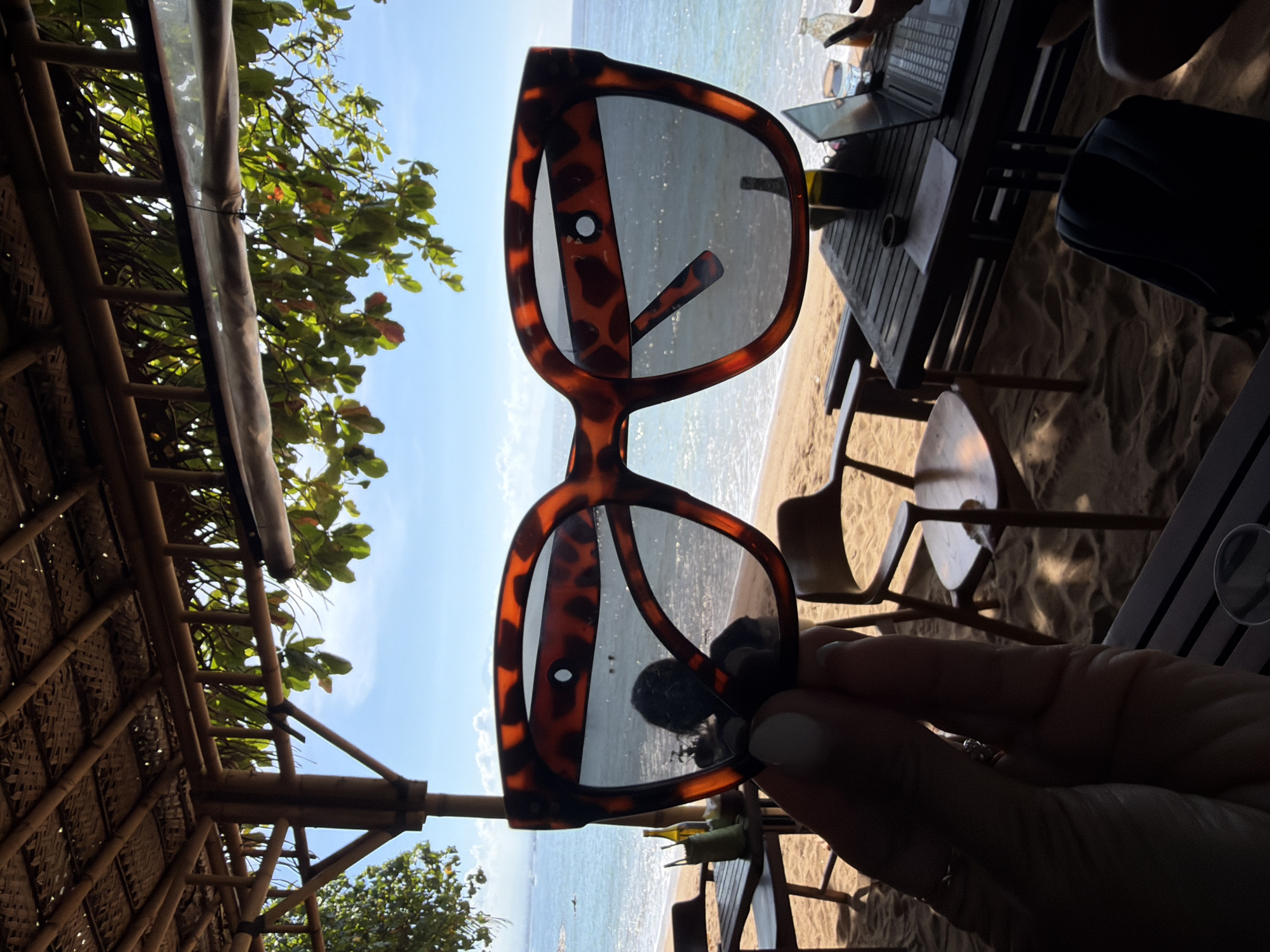 This image is a close-up of a person's hand holding a pair of tortoise-shell sunglasses in the foreground. The sunglasses are held up, obscuring a portion of the background scene. The background shows a beach with sand, several wooden tables and chairs, and a thatched-roof structure providing shade. Beyond the beach is the ocean with calm water and a clear blue sky with some wispy clouds. There are some trees with green leaves casting shadows on the sand. A laptop, a glass of water, and a bottle are visible on one of the tables. The overall scene suggests a relaxed, tropical setting, likely a beachside cafe or restaurant in Denpasar, Indonesia.