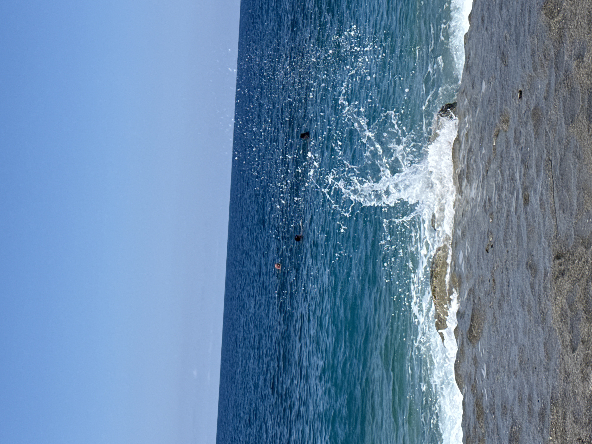 The image shows a coastline on a clear day. The sky is a solid blue. The sea is a deep blue color, and there are three people swimming in the water. The sea is choppy, with many white waves. The coastline is rocky and gray. The image is rotated 90 degrees.