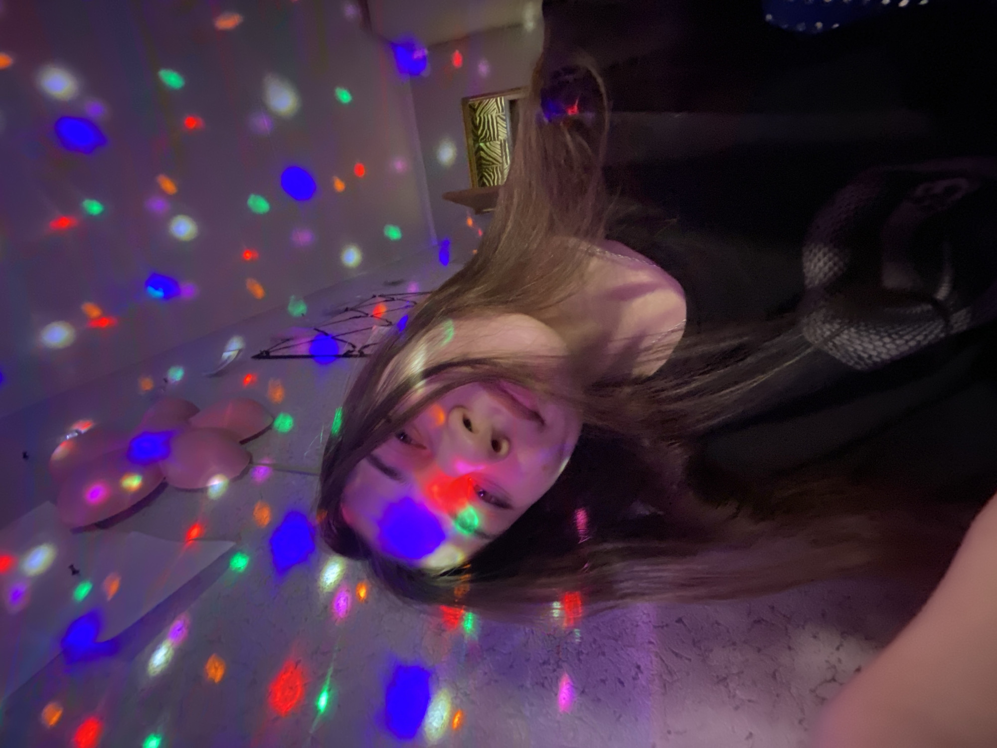 A young woman with long, brown hair is photographed indoors under colorful, patterned lighting. Her face is illuminated by blue, red, green, and other colored spots, suggesting a disco ball or similar lighting effect. The background includes a white wall with more of these light patterns, a bed with pink balloons. No evidence indicates this image was taken in Votkinsk, Russia.