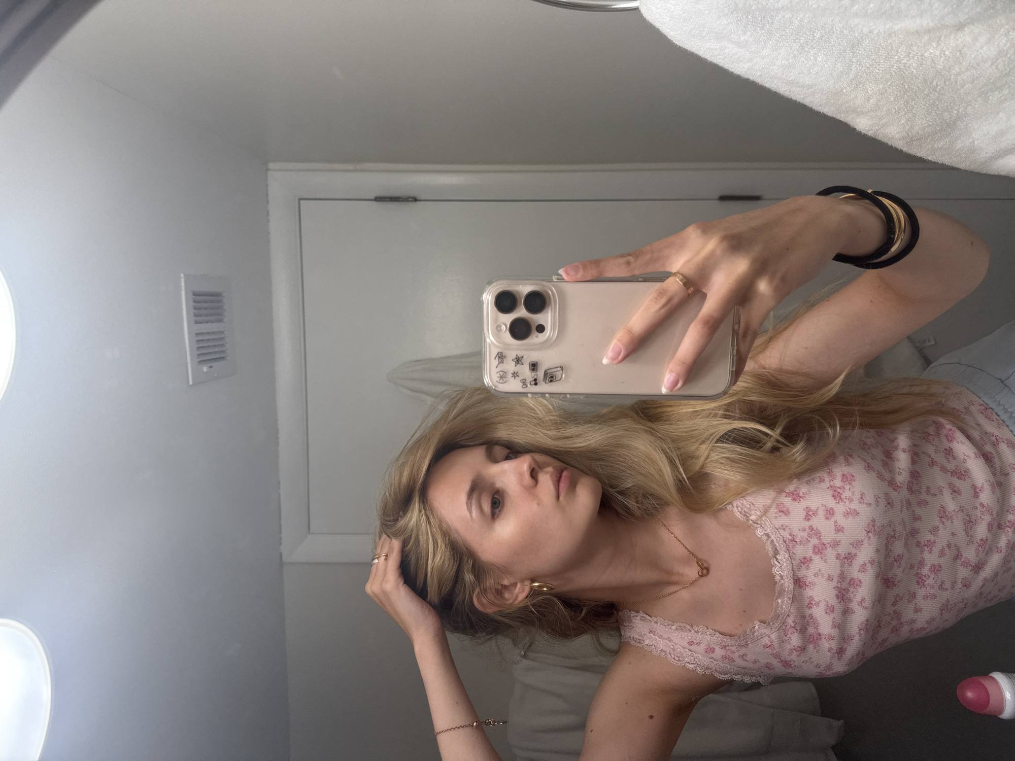 A young Caucasian woman with long blonde hair is taking a mirror selfie in what appears to be a bathroom. She is wearing a pink floral tank top with lace trim and several pieces of gold jewelry, including hoop earrings, a necklace, and rings. She is holding her phone, which has a clear case with stickers, in her right hand and touching her hair with her left hand. A white towel hangs to the right of the frame, and a white door is visible in the background. The environment is well-lit, with ceiling lights visible at the top of the image. There is a vent on the wall above the door. No specific Miami landmarks or indications of the city are present in the image.