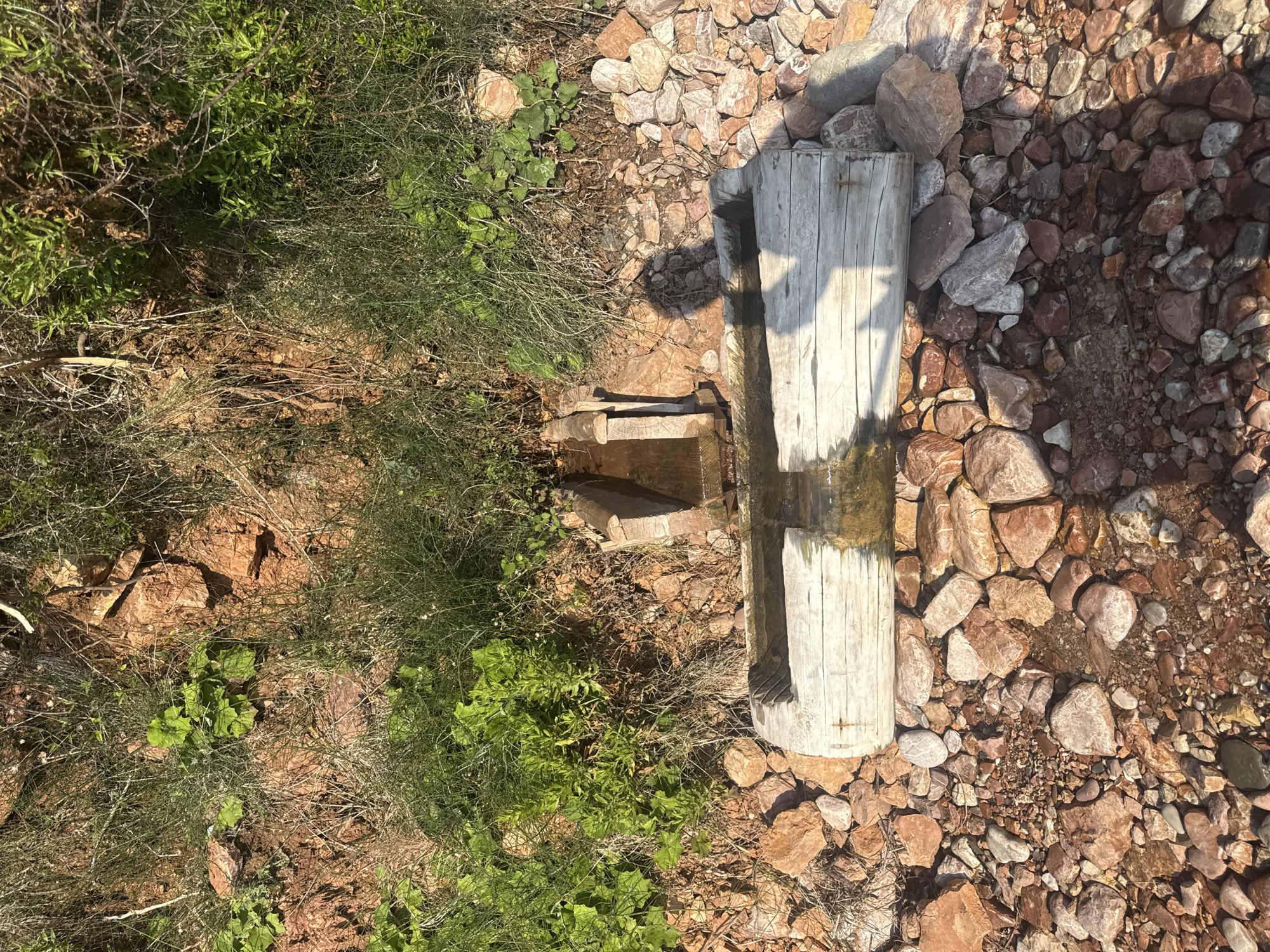 The image shows a natural spring flowing into a carved wooden trough, located in Sveti Stefan, Montenegro. The trough is situated on a rocky embankment covered with sparse vegetation. The water is clear and reflects the sunlight. The surrounding area is characterized by reddish-brown soil and various sizes of rocks. The lighting suggests it is daytime, with shadows cast by the trough and surrounding elements.