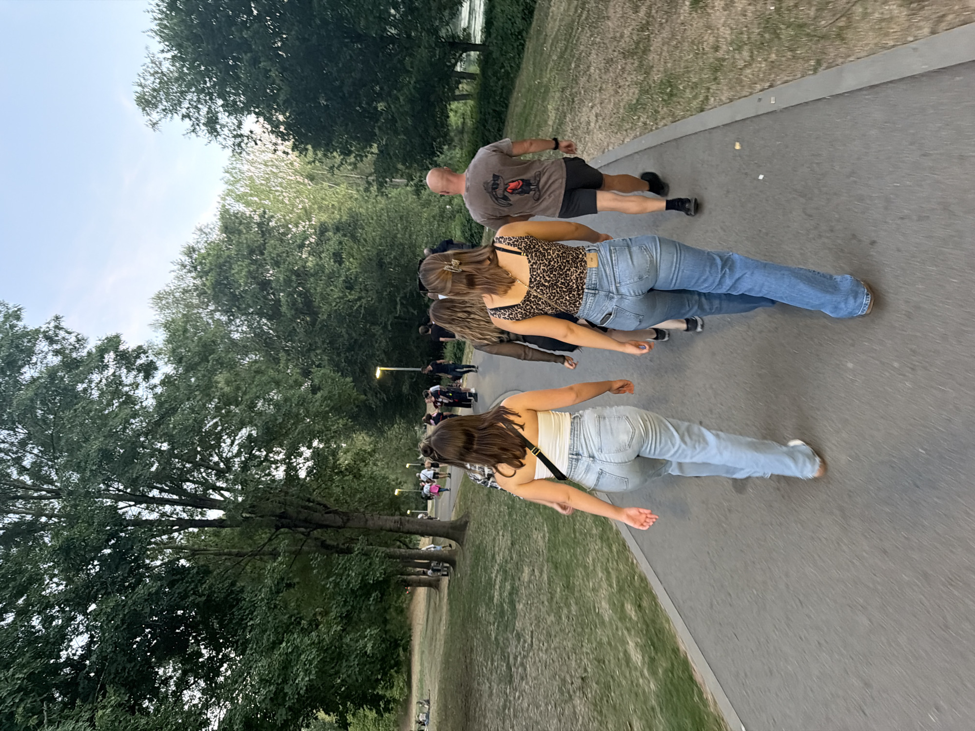 In Echternach, Luxembourg, a group of people are walking along a paved path in a park. In the foreground, two women and a bald man are walking away from the camera. The woman on the left is wearing a white strapless top and light blue jeans. The woman in the middle is wearing a leopard-print halter top and light blue jeans. The man on the right is wearing a grey t-shirt with a graphic print and dark shorts. Behind them, other individuals are visible walking further down the path, and some people are seated on the grass to the left. The park features mature trees with dense foliage on either side of the path, and the sky above is a pale blue with some wispy clouds. The ground is covered with dry grass and scattered cigarette butts.
