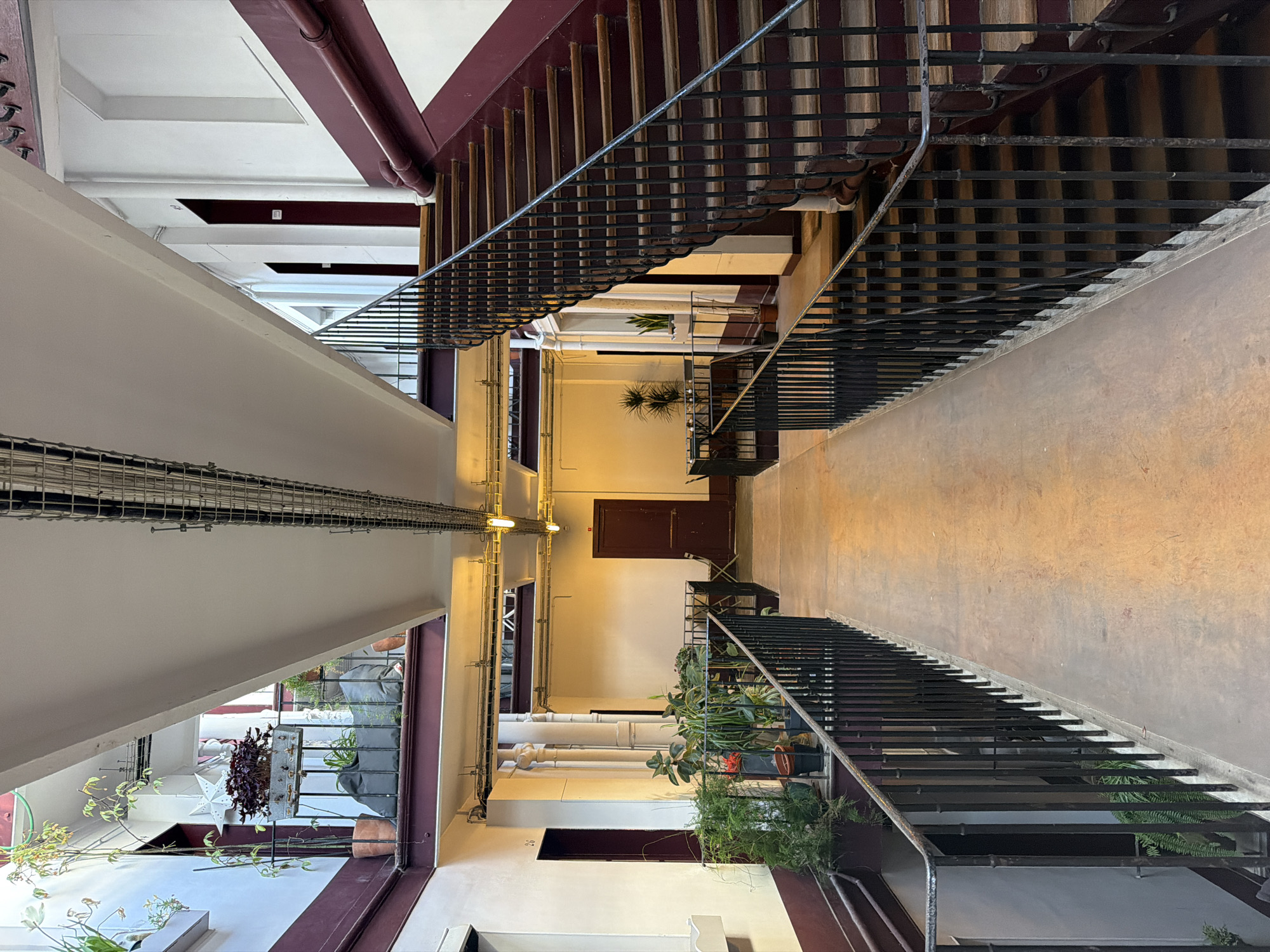 The image shows an interior view of an atrium, likely in Paris, France, given the architectural style. The scene is captured from a relatively high angle, looking down the length of the space. Two flights of stairs with black metal railings flank the central area. The staircases are constructed with wooden treads and burgundy-painted support structures.
The walls are a pale yellow, with visible conduits and pipes running along them. Several doorways are visible on each floor, painted in a matching burgundy color. Potted plants and greenery are placed along the railings and windowsills, adding life and color to the scene.
The ceiling features exposed beams, and a metal grid system is suspended from it. The overall impression is of a functional, older building with some attempts at interior decoration. The scene is devoid of people.