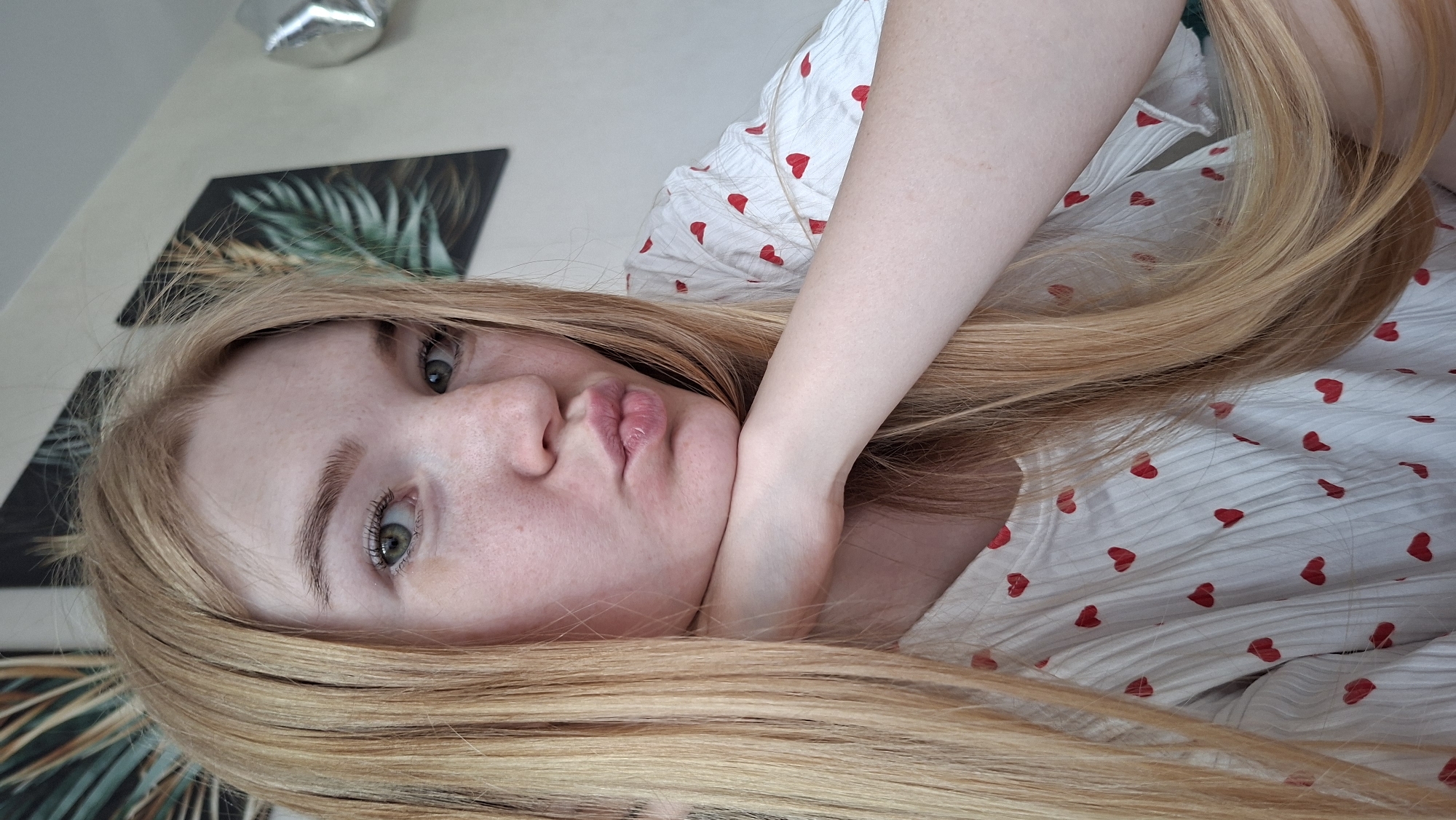 A fair-skinned woman with light blonde hair is lying down, facing the camera. Her left arm is bent and positioned with her hand under her chin, creating a somewhat pouty expression. She has freckles across her nose and cheeks. She is wearing a white top or dress with small red hearts patterned across it. In the background, there is a decorative artwork with a botanical design, and a silver balloon. The specific location within the city of Chaikovsky, Russia, cannot be determined from the image alone.