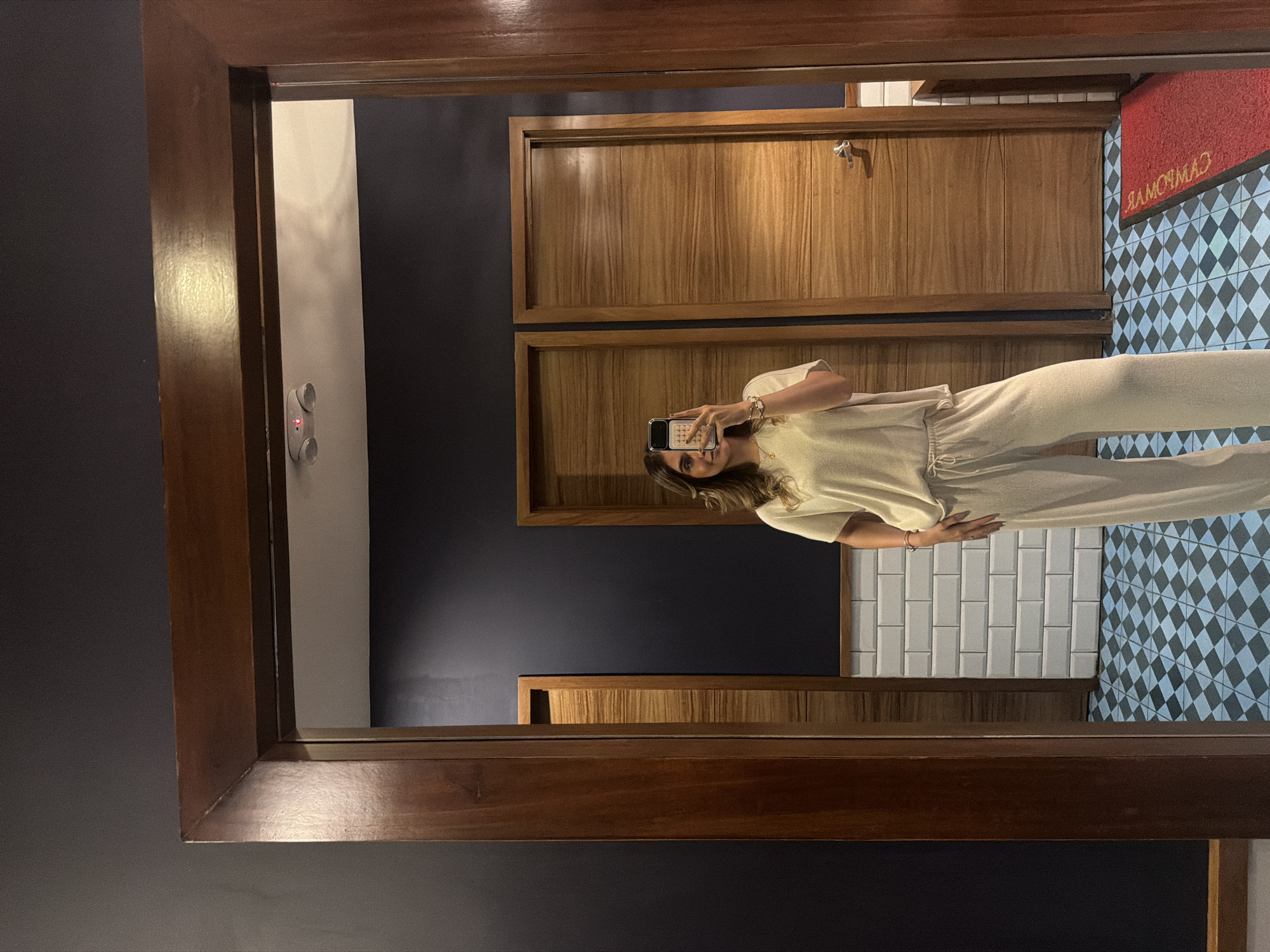 A mirror reflects a woman holding a smartphone, presumably taking a selfie, in a room with a contemporary design. The woman is wearing a light-colored two-piece outfit. The background features dark blue walls, wooden cabinetry, white subway tile walls, and a patterned tile floor in black and light blue. The mirror has a wooden frame. A red mat with white lettering is visible on the tiled floor. The scene is in a bathroom, likely located in Puebla, Mexico, as suggested by the lettering on the mat which reads backwards as "CAMAROMAD," potentially referring to "Camaro Mad," a local brand or location.