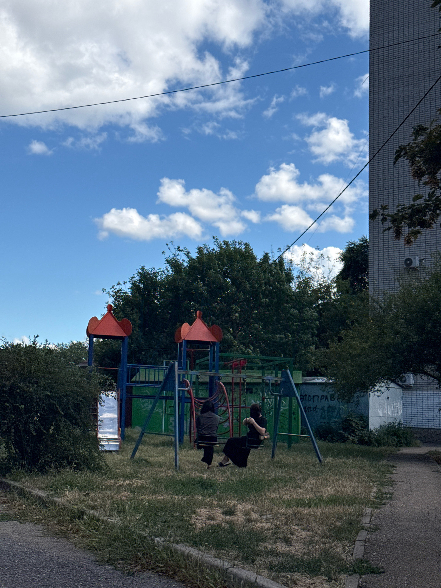 An outdoor, eye-level, full shot shows a playground on a cloudy day in an urban setting. In the foreground, a paved area transitions to a grassy lawn. A playground structure with red and blue metal components is visible, as well as a green metal fence behind it. Two people, dressed in black, are seated on swings. The person on the left is facing away from the viewer, and the person on the right is facing towards the viewer. A tall, grey brick building with windows is to the right of the playground. Trees and bushes surround the playground area. Power lines are strung across the sky. A reflective surface, possibly a mirror, is leaning against a bush on the left side of the frame. Graffiti is present on a white surface behind the playground structure.