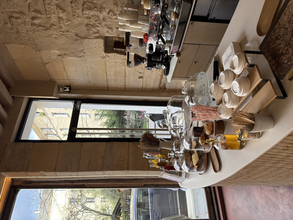 The image depicts the interior of a cafe in Paris, France, viewed from a slightly elevated perspective towards a service counter and large windows. The central focus is a light-colored, curved counter with a textured, ribbed facade.
On the counter, from left to right: a collection of bottled beverages in various hues (yellow, orange, red) stands beside a white pitcher holding dried flora. Several glass cake stands with domed covers display an assortment of baked goods, including cakes and pastries such as canelés. Adjacent is a tall glass filled with red and white striped paper straws. Further right, a clear dispenser bottle with a golden pump contains a yellowish liquid. A ceramic pot holds wooden stirrers. Stacks of white paper cups with matching lids are arranged on a tiered wooden display stand. White napkins, one prominently displaying "MOR," are placed nearby. In the foreground on the right, a large, antique-style book with a dark, textured cover and a small wooden tray are visible.
The back wall of the cafe features exposed, light-colored hewn stone blocks. A recessed niche within the stone wall contains a few small objects. A professional-grade espresso machine, silver and black, with multiple group heads and steam wands, is situated on a counter beneath the stone wall, accompanied by a coffee grinder filled with beans. Various cups are stacked on the espresso machine and surrounding counter space. Two white security cameras are mounted high on the wall, above the window frame. Wooden ceiling beams are visible at the top of the frame, and a reddish-brown, textured floor is seen in the lower left.
Large, dark-framed windows dominate the left and center-right of the background, offering views to the exterior. Through the left window, a street scene is visible with a dark-colored car parked, bare deciduous trees, and a light-colored building in the distance. The right window reveals a small courtyard or side street, featuring a red bicycle parked near green bushes and trees, with another light-colored building beyond. The sky is bright and clear. No individuals are present within the scene.
