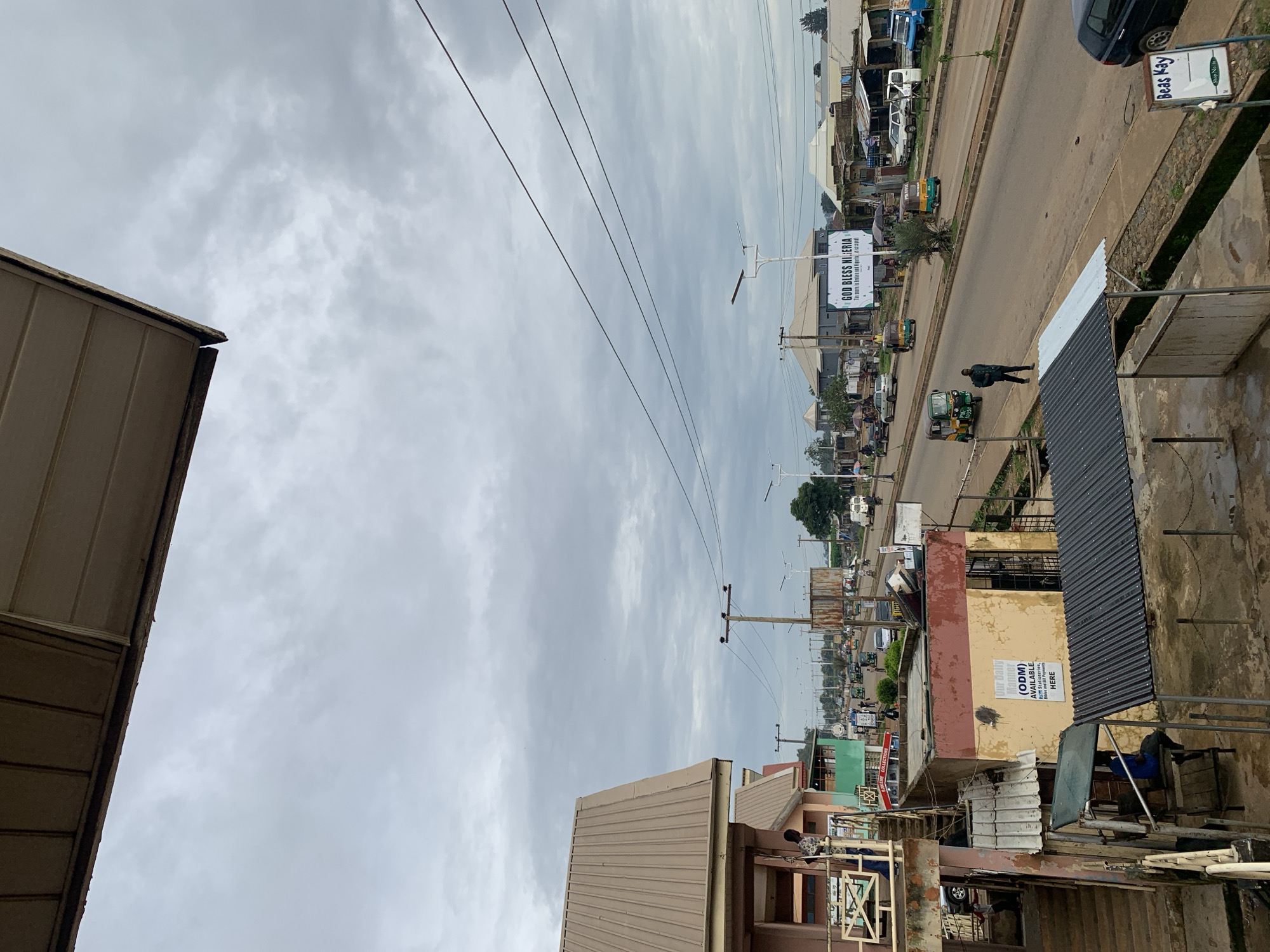 The image depicts a street scene in Rafin Par, Nigeria, under an overcast sky. The foreground features the side of a building with a corrugated metal roof and some external stairs. A person is visible standing on a balcony-like structure. Further down the street, a road is lined with various buildings, shops, and stalls. Several vehicles, including cars, tricycles (keke napep), and a van, are present on the road. People are seen walking and some are under umbrellas, suggesting the possibility of light rain. A large banner with the text "GOD BLESS NIGERIA" is prominently displayed on the side of a building. Utility poles with electrical wires run along the street. The overall impression is of a busy urban environment, likely a market area or a commercial district in a Nigerian town.