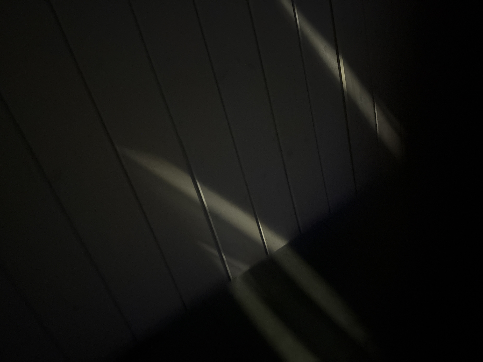 The image depicts a close-up, abstract view of a dark, textured surface, possibly wood paneling or siding, with diagonal light streaks. The lighting creates a dramatic effect, with strong contrasts between illuminated and shadowed areas. There are no people, specific objects, or identifiable environmental features in the scene. The image does not provide any information about its location in Doma 1096 Km, Russia.