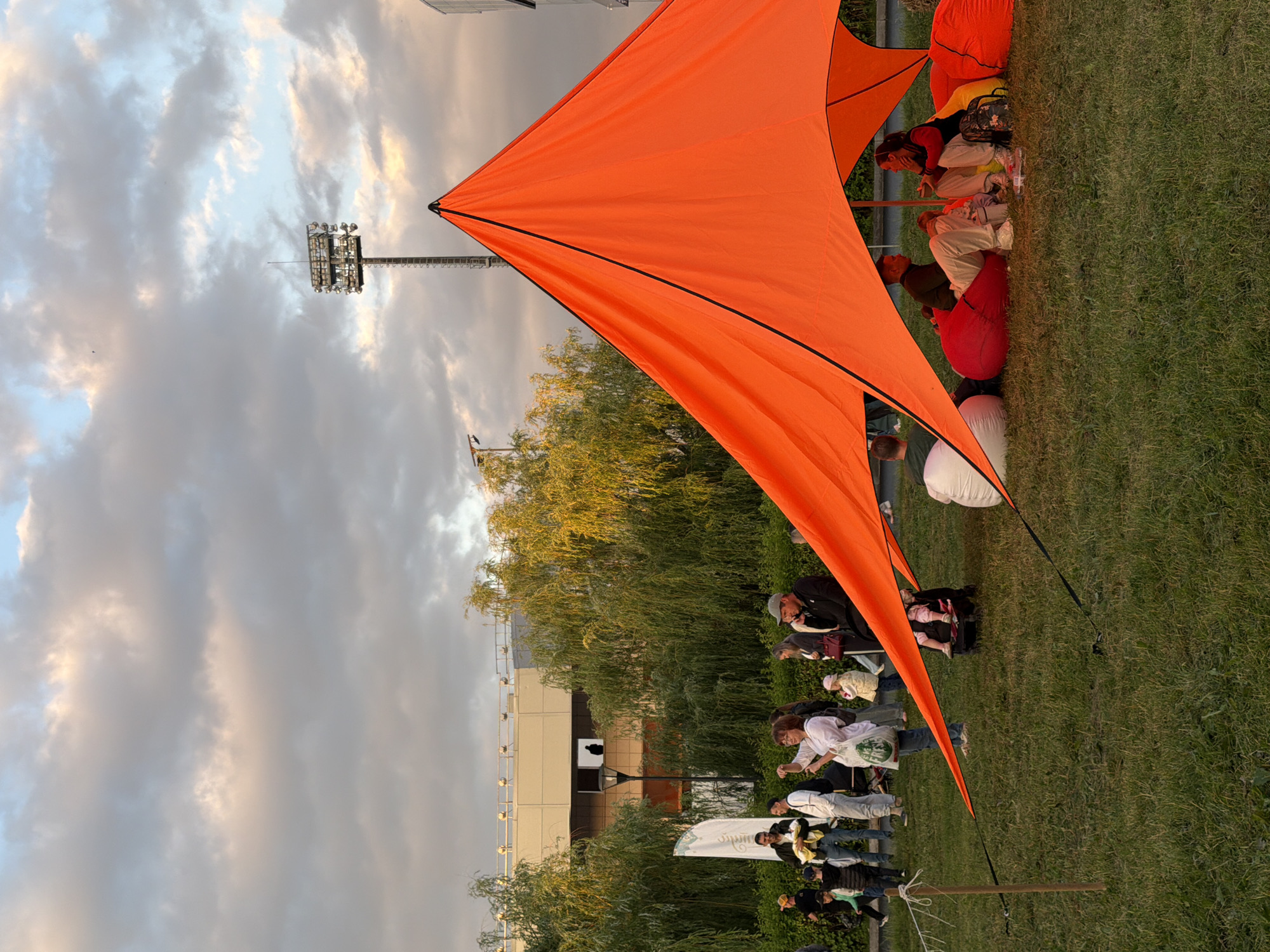 The image depicts an outdoor gathering with an orange tent as the central focus. Several people are present, some standing and observing, while others are seated on beanbag chairs under the tent. In the background, there are trees, a building with windows, and a tall stadium light. The sky is filled with clouds, suggesting it may be late afternoon or early evening. The ground is covered in grass.