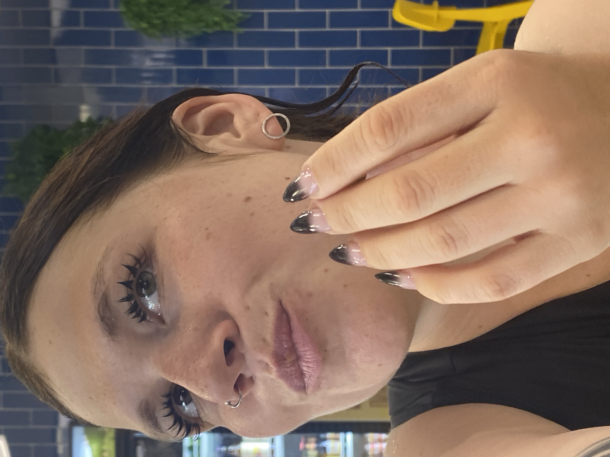 The image shows a close-up of a woman's face, with a particular focus on her eyes, nose, mouth, and hand. She has dark brown hair, light-colored eyes, and visible freckles on her skin. Her eyelashes appear to be enhanced. She is wearing a nose ring and a circular earring. Her fingernails are painted with a gradient of clear polish at the base, transitioning to black at the tips. Her left hand is raised towards her face, with her fingers touching her cheek. She is wearing a dark-colored tank top. The background consists of blue tiled walls and a blurry yellow object, possibly a chair, in the distance. There are also hints of greenery and shelves with bottles. The scene is indoors, likely in a public place. There is no information or visual cues to indicate the location is Votkinsk, Russia.