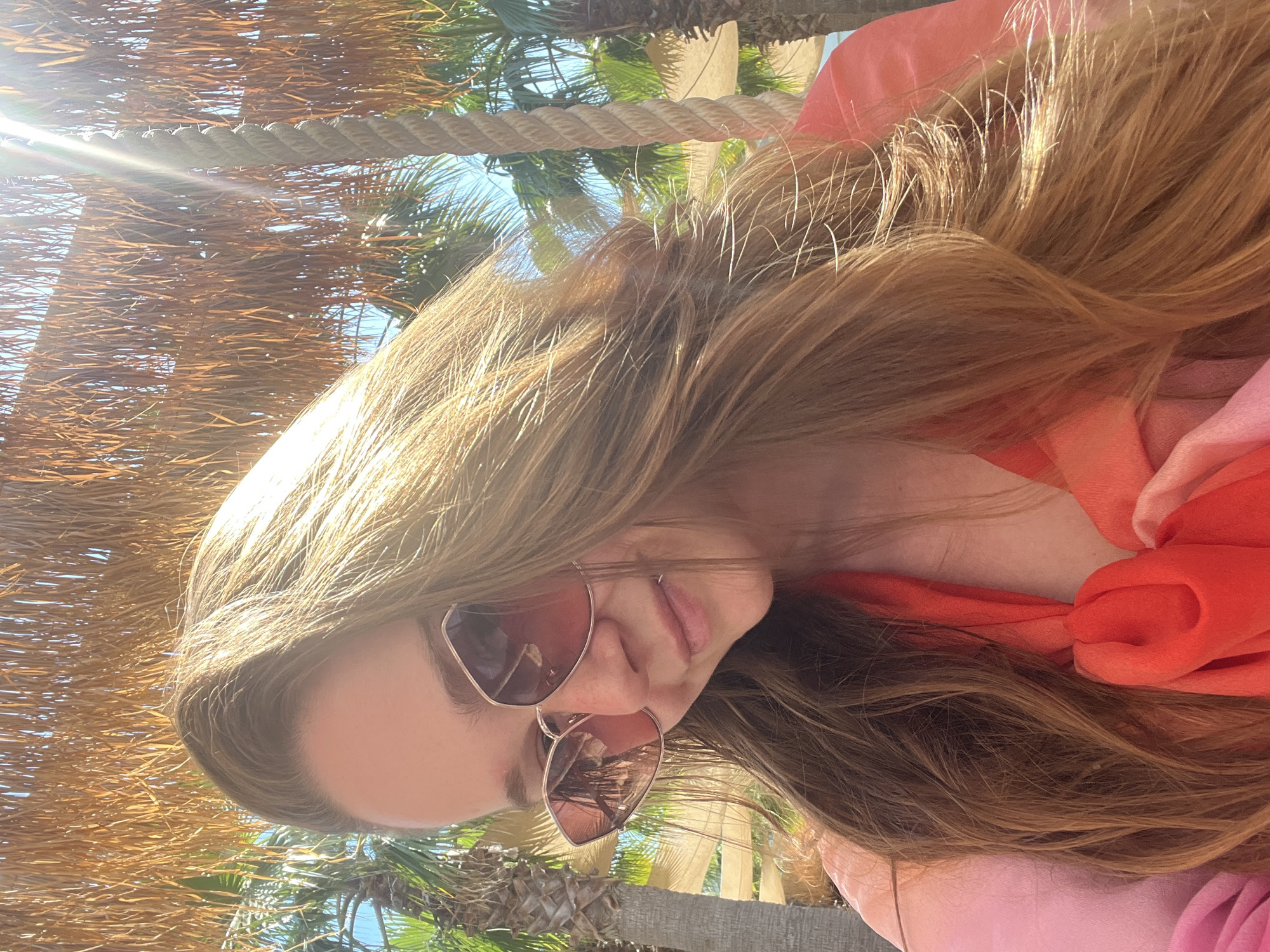A medium shot taken outdoors depicts a young woman with long, light brown hair wearing oversized pink-tinted sunglasses. She is looking downwards and to her left, with a slight smile. She is wearing a bright pink and orange garment, possibly a dress or scarf, draped around her shoulders. The background features a thatched roof structure, palm trees, and a clear blue sky, suggesting a sunny, tropical or Mediterranean environment. The overall lighting indicates a bright, sunny day, with some sun flare visible. The image does not provide explicit details about the city of Thassos, Greece, but the environment is consistent with a coastal resort area.