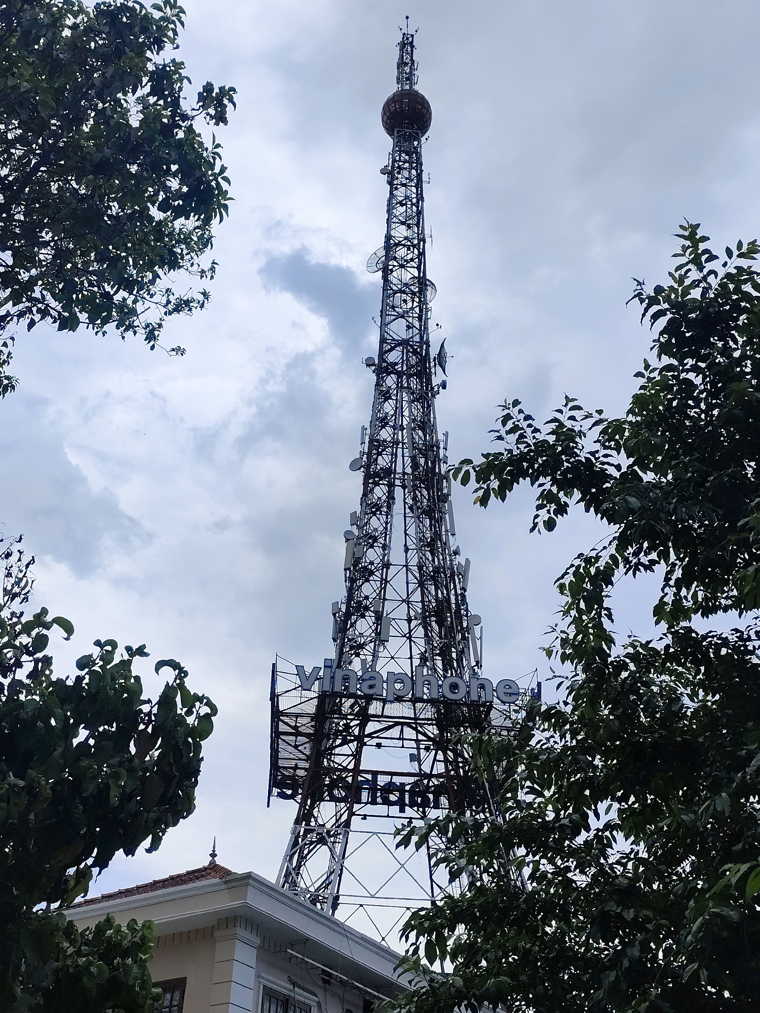 This is a low-angle, full-shot photograph of a telecommunications tower in Da Lat, Vietnam, framed by trees on both sides. The tower, a tall, lattice-structure with numerous antennas and satellite dishes, is the central subject. The word "vinaphone" is prominently displayed in large, blue letters on a rectangular signboard attached to the tower. The sky above is cloudy with patches of blue peeking through. In the lower left corner, the roof and upper portion of a light-colored building with a tiled roof are visible. The overall impression is one of industrial infrastructure set against a natural and urban backdrop.