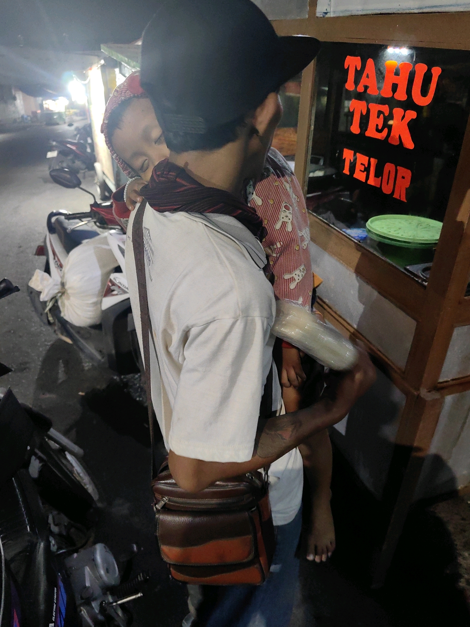 In Keboansikep, Indonesia, a person wearing a black cap and a light-colored t-shirt is carrying a sleeping child. The child is wearing a red and white striped outfit with bunny motifs and a red beanie. The person has a brown and tan shoulder bag. In the background, a sign with red lettering reads "TAHU TEK TELOR." Several motorcycles are parked nearby, and a green plastic bowl is visible on a counter. The scene appears to be outdoors at night, possibly near a food stall.