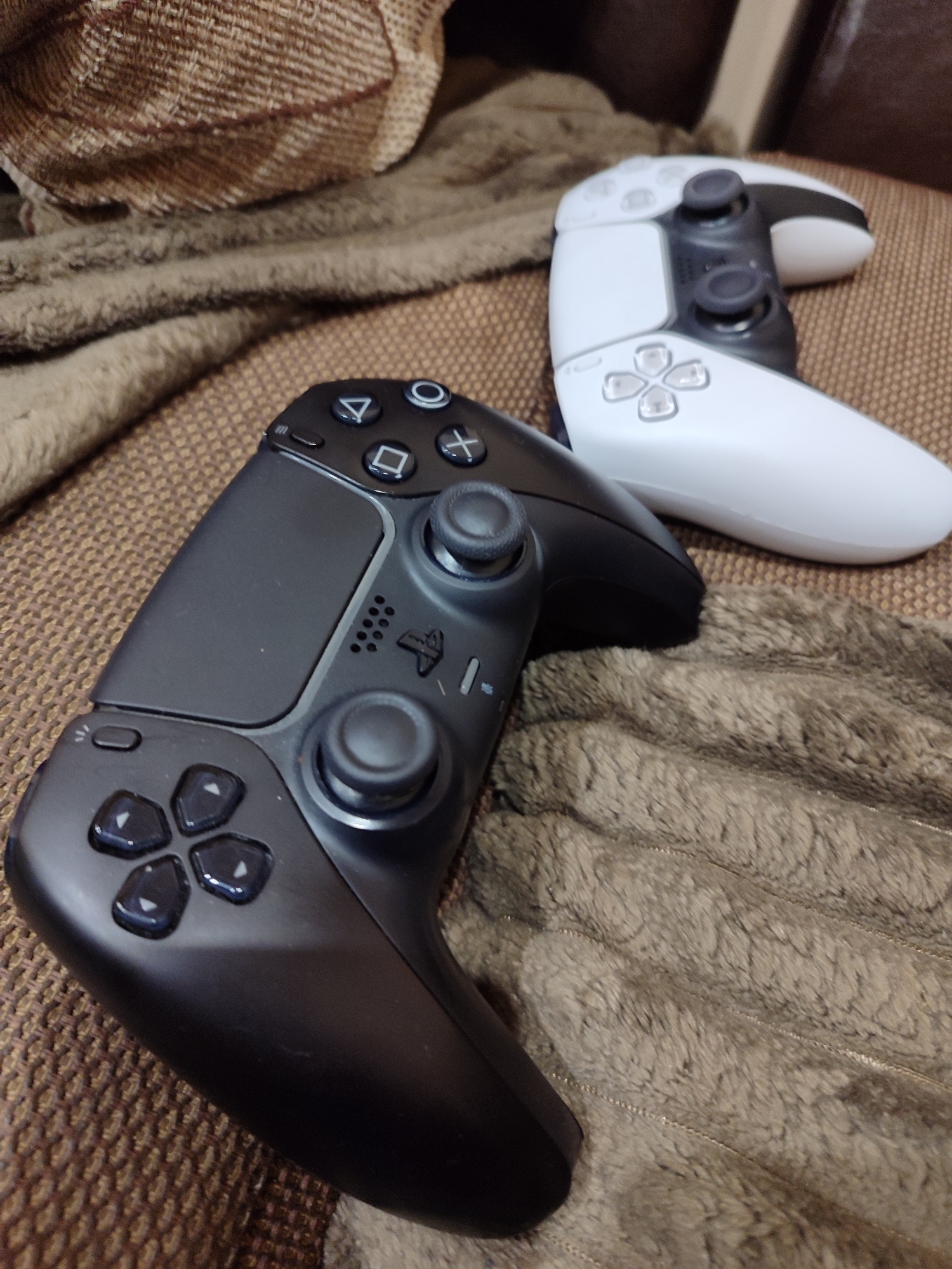The image shows two PlayStation 5 DualSense controllers resting on a textured surface, possibly a throw blanket or upholstery. One controller is black, positioned in the foreground, angled towards the viewer. The second controller is white, placed behind the black one. The background features more of the textured surface and indistinct objects. There is no indication of Burgas, Bulgaria in the image.