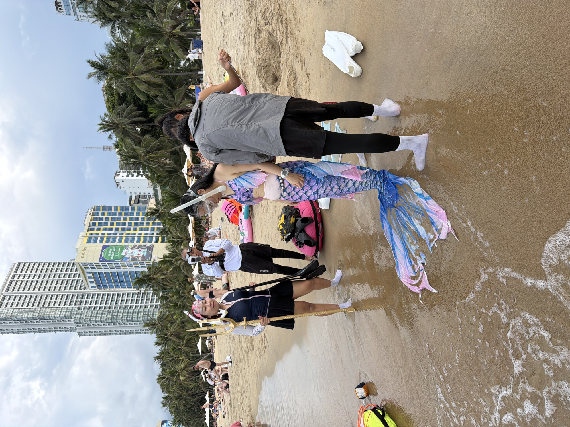 The image depicts a beach scene in Nha Trang Ward, Vietnam, with several people present. The primary subjects are two individuals in what appears to be a costume or performance. One person is dressed as a mermaid, wearing a colorful tail with shades of blue and pink, and a matching top. They are also wearing a snorkeling mask and holding a golden trident. The other person is dressed in a dark blue swimsuit with a skirt and also wears a pink headband and goggles, holding a similar trident. They are standing on the wet sand near the water's edge.
In the background, other individuals are visible on the beach, some seated, suggesting a gathering or event. Behind the beach, tall buildings and palm trees are prominent, indicating an urban coastal setting. The sky is cloudy, hinting at overcast weather. The presence of snorkeling gear and mermaid attire suggests a recreational activity or a themed event, possibly a photo shoot or performance. The overall mood appears casual and festive.
Several objects are scattered on the sand, including white fins, a pink inflatable, and a yellow life vest. The water is gently lapping onto the shore, creating small waves and foam. The lighting suggests it is daytime. There is some visible text on a billboard-like sign on one of the buildings, but it is largely illegible. The image's orientation is rotated, with the subjects appearing to stand vertically on the sand.
