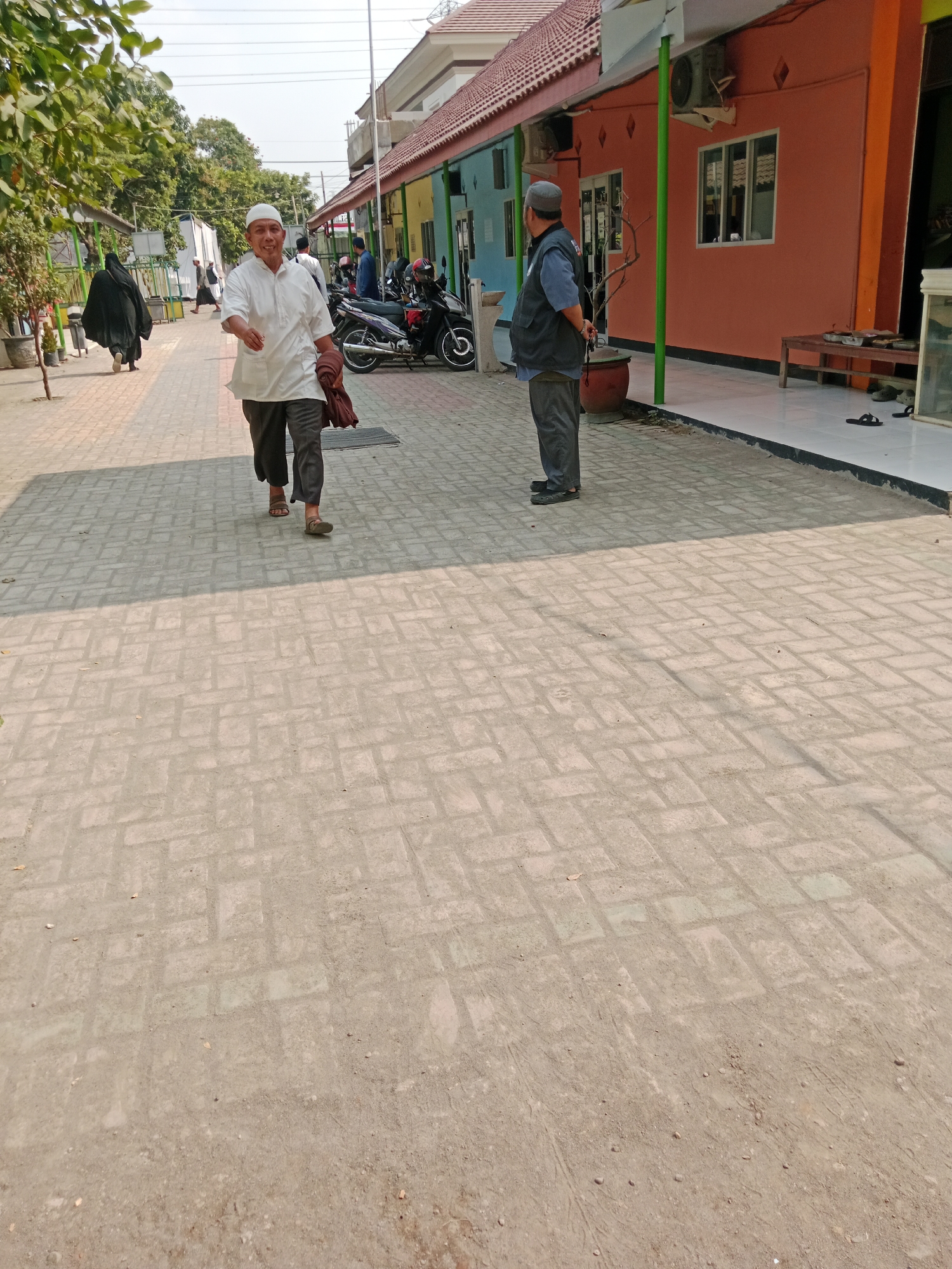 In Kediri City, Indonesia, an outdoor scene unfolds along a walkway paved with light-colored, rectangular bricks arranged in a basketweave pattern. The walkway is bordered by buildings with brightly colored facades – shades of coral, light blue, and orange – and a tiled, white entryway. Green support poles stand in front of the buildings. On the walkway, a man wearing a white shirt, grey pants, a white skullcap, and sandals is walking towards the viewer, holding a brown cloth over his right shoulder. He is smiling. Further down the walkway, another man in a dark vest, blue shirt, grey pants, and a grey skullcap stands facing away from the camera, his hands behind his back. Several motorcycles are parked near the buildings. In the background, trees with green foliage are visible, and a clear, bright sky suggests it is daytime. The overall impression is one of a bright, sunny day in a possibly religious or community setting.