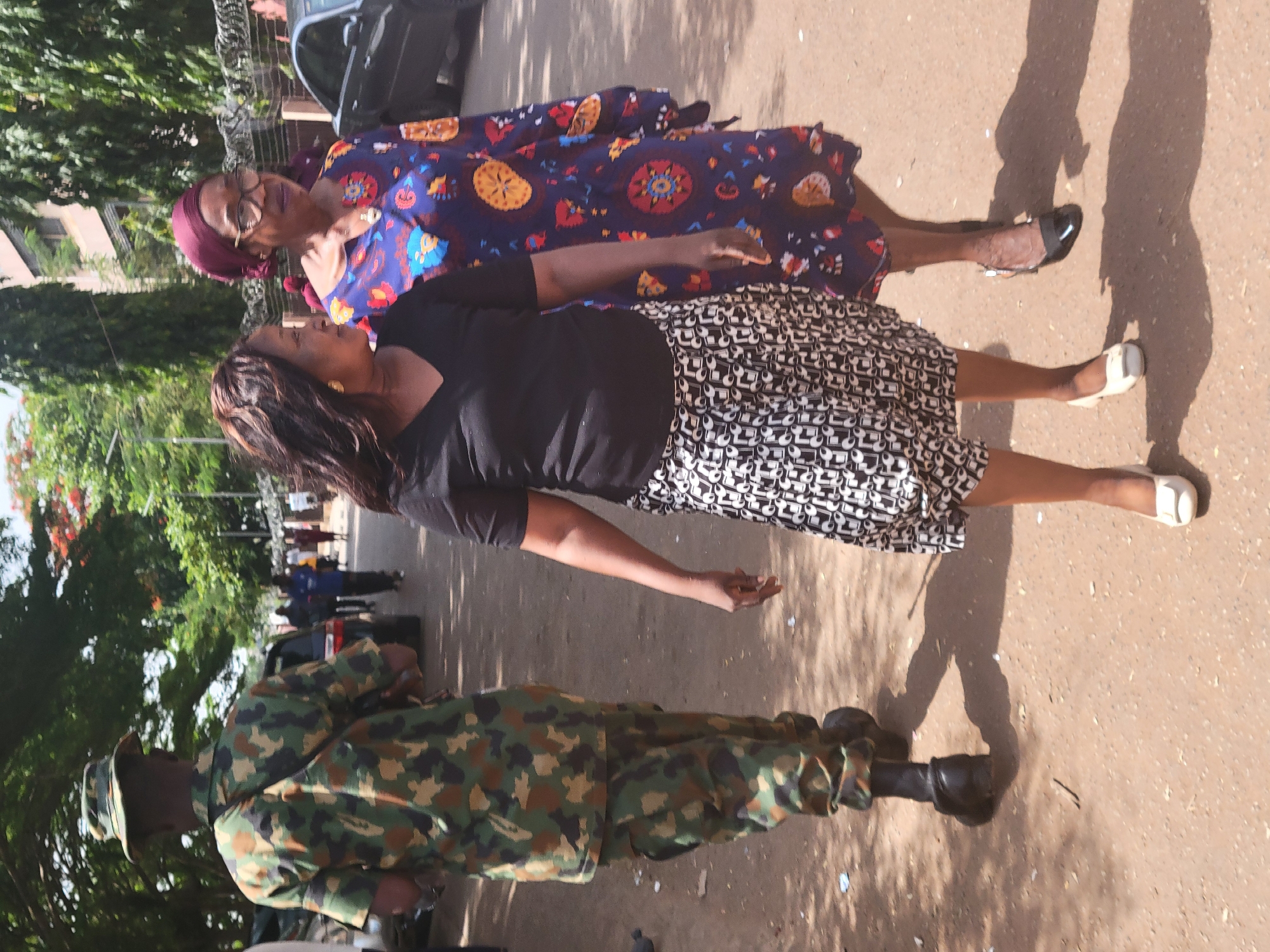 The image, taken in Abuja, Nigeria, captures an outdoor scene featuring three individuals under bright daylight. In the foreground, seen from behind, is a person wearing a full camouflage military uniform, including a matching hat. Their shadow is distinctly cast on the light-colored ground.
To the right of the military personnel, in the middle ground, stand two women. The woman closer to the foreground has dark, long hair and is dressed in a black short-sleeved top paired with a black and white patterned knee-length skirt. She wears light-colored flat shoes, and her right arm is slightly extended to her side.
Behind her, the second woman is visible wearing glasses and a maroon headscarf. She is adorned in a vibrant, loose-fitting blue dress patterned with large red, orange, and yellow circular designs, and wears black flat shoes. Both women are looking towards the left side of the frame, appearing calm.
The background features lush green trees and foliage, indicating a natural or landscaped environment. A dark vehicle is partially visible behind the women. Strong shadows on the ground signify bright sunshine, suggesting it is daytime. The presence of military personnel alongside civilians points to a public or semi-public setting. There is no visible text in the image.