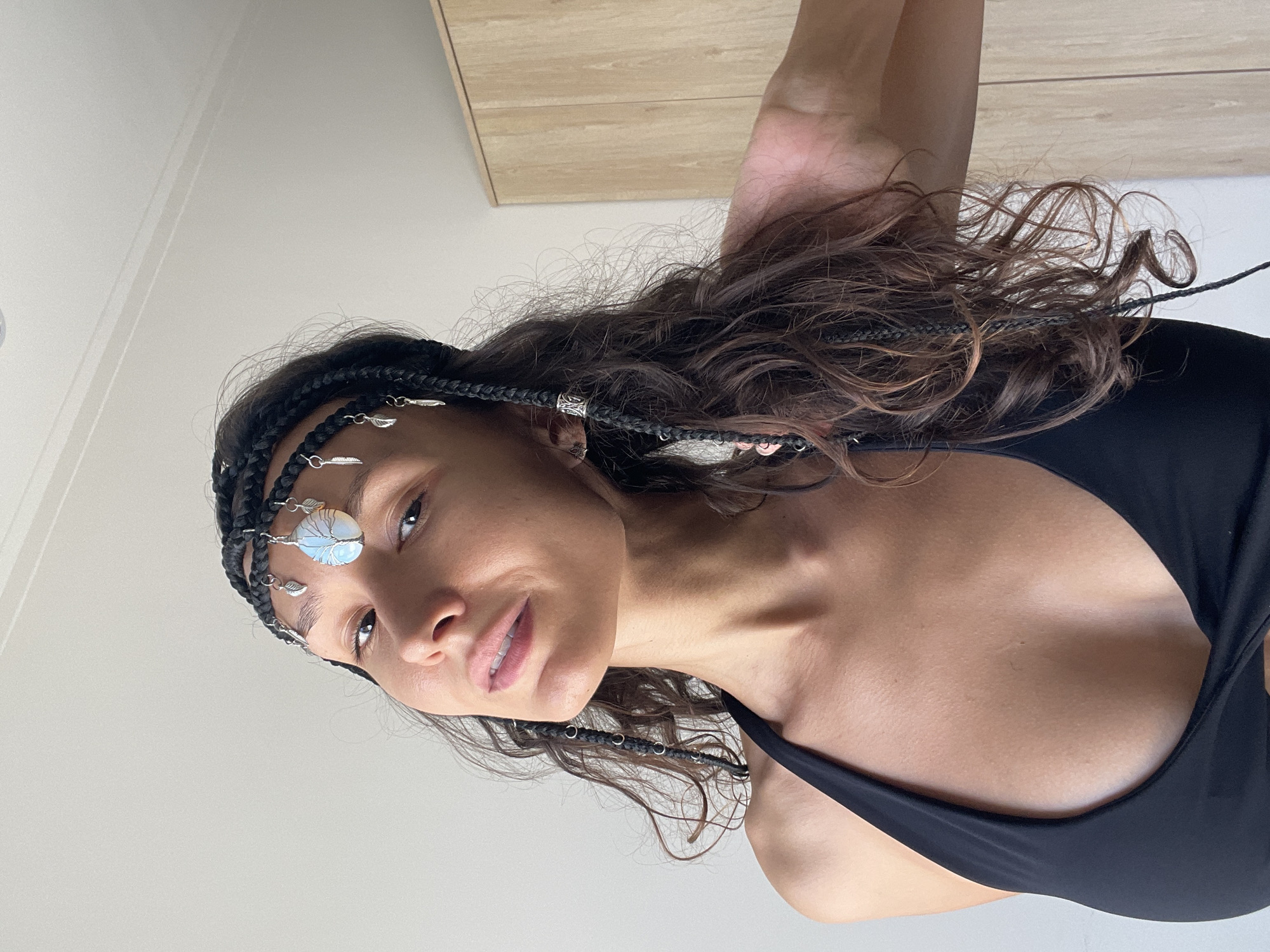 A young woman with dark hair styled in braids and loose curls wears a black halter top and a braided headband adorned with a tree of life pendant and small silver charms. She is looking at the camera with a slight smile. The background appears to be an indoor setting with a plain wall and a wooden wardrobe. The lighting is natural, casting soft shadows. No specific details suggest the location is Phu Thuy Ward, Vietnam.