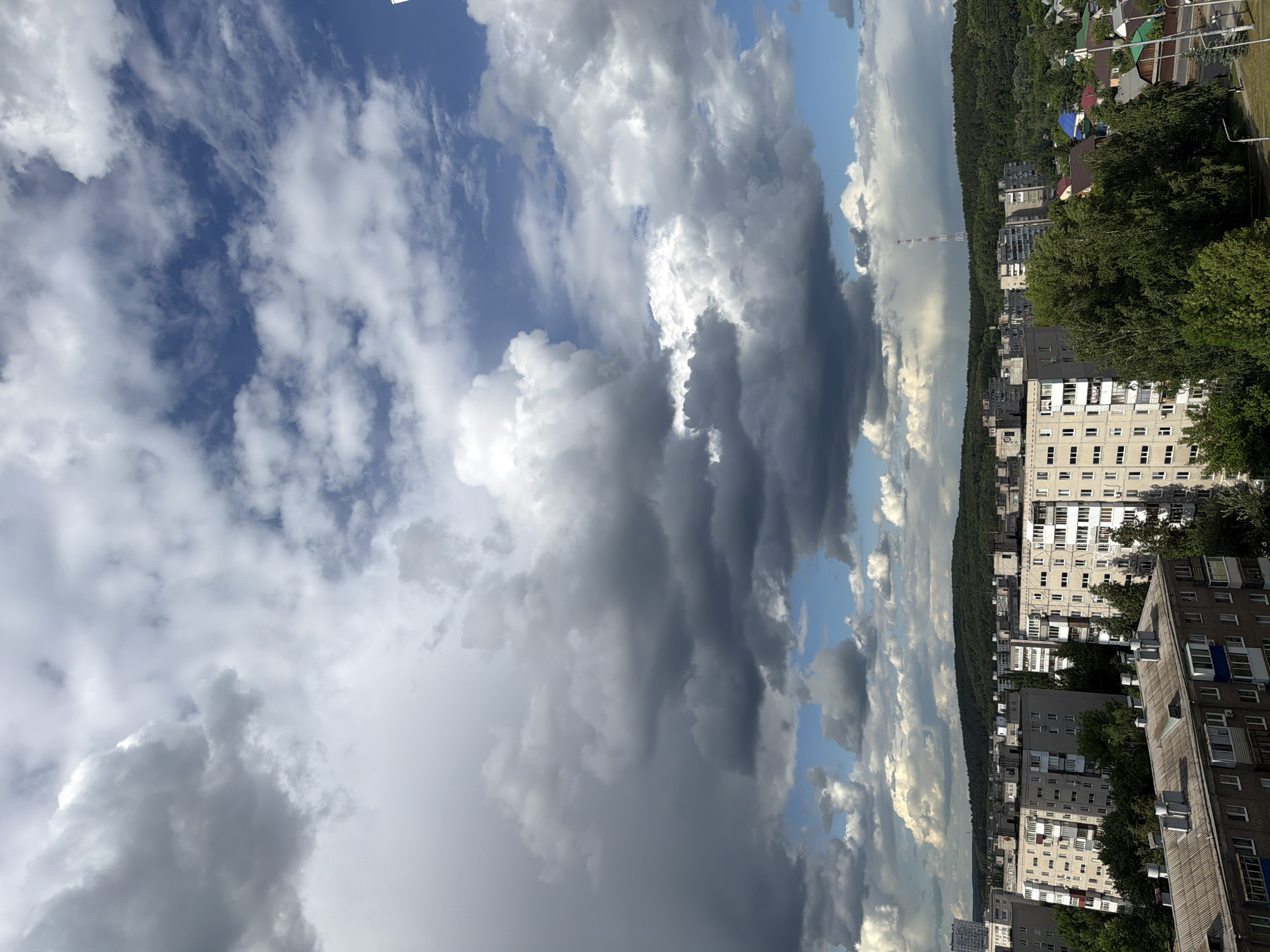 The image shows a view of a city with a sky filled with clouds. A mix of light and dark clouds fills the upper portion of the image, with patches of blue sky visible. In the lower portion of the image are several apartment buildings arranged along the horizon with a green forested area behind. A tall metal communication tower is visible between the buildings. Some houses with colored roofs can also be seen among the trees.