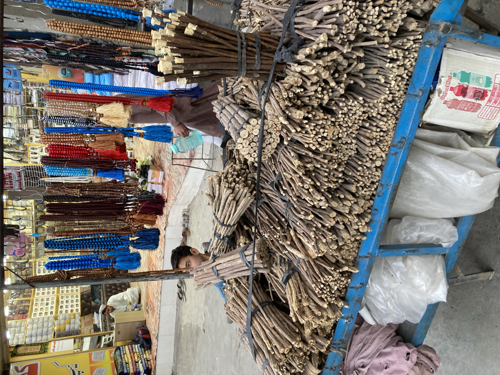 This image captures a vibrant market stall scene in Quetta City Tehsil, Pakistan, bustling with activity and goods. The perspective is slightly angled, looking down into the market space.
In the foreground, a prominent blue metal cart on the right is heavily loaded with numerous bundles of wooden sticks, likely miswak (chewing sticks used for dental hygiene). These dark brown sticks are tightly bound with black straps, and many bundles feature lighter, possibly prepared or chewed ends. On the lower shelves of this cart, several white plastic bags and a cardboard box are visible, with the box showing partial text "500 ml. Ped" alongside illustrations of beverage bottles. The face of a young boy is partially visible, peeking out from behind one of the stick bundles in the mid-right.
To the left, a brightly colored display rack showcases a wide array of prayer beads (tasbih or misbaha) in various colors, including shades of blue, red, brown, black, white, and gold, many adorned with matching tassels. In the mid-ground, a person wearing a brown traditional garment is visible, holding a light blue bottle. Further in the background, another individual in a white garment is seen kneeling or sitting amidst shelves stocked with numerous small, packaged items, suggesting an active retail setting.
The ground appears to be a tiled or patterned floor. The lighting indicates it is daytime. The scene conveys the busy atmosphere of a traditional street market or bazaar, with an emphasis on local products such as miswak and prayer beads, reflecting the cultural context of the region. No strong emotions are discernible from the individuals, who appear engaged in typical market activities.