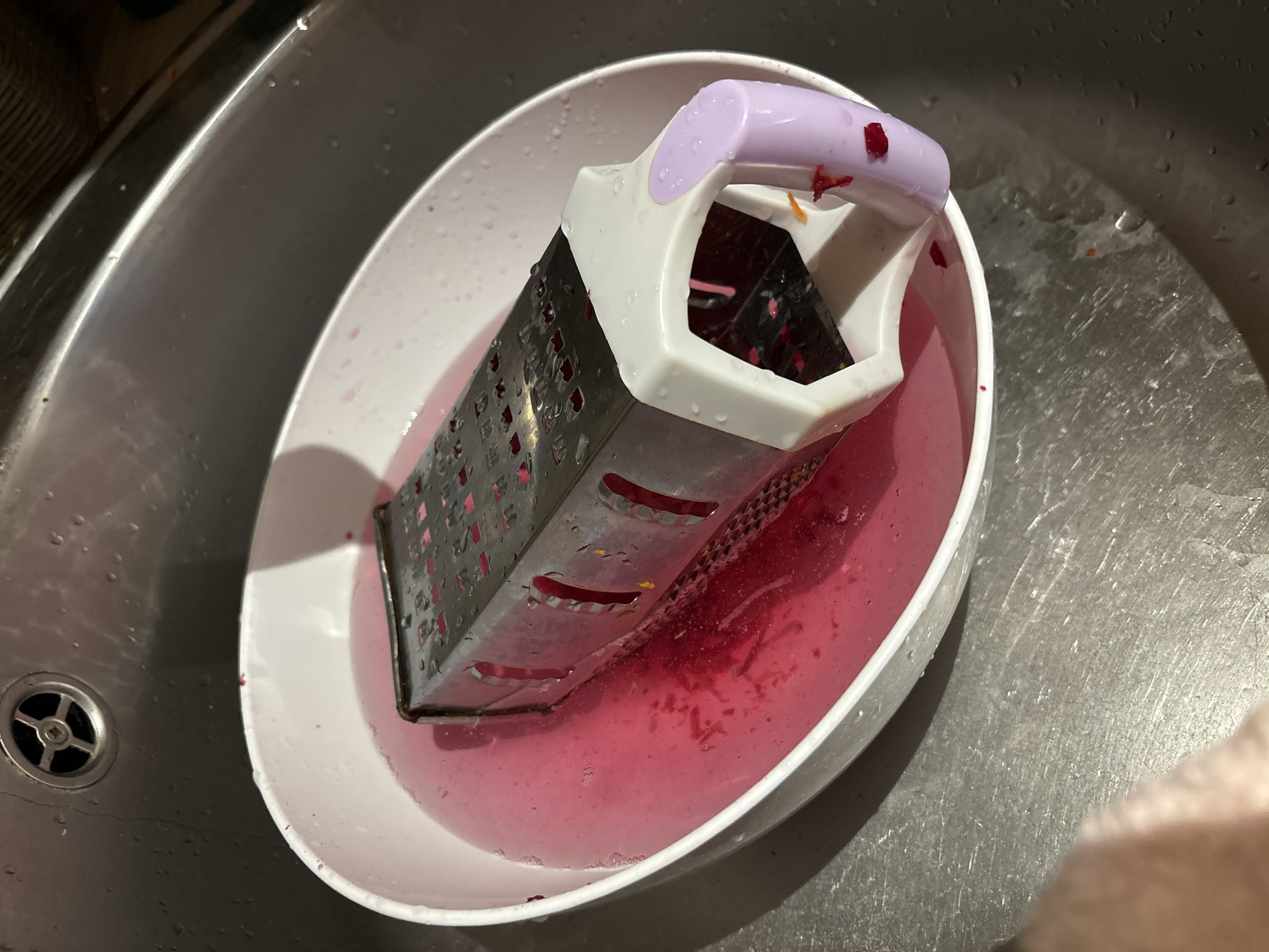 A grater sits in a white bowl filled with pink liquid, presumably water and beet juice, in a stainless steel sink. Small pieces of what appears to be beetroot are visible in the liquid and on the grater. The grater has a metal body with various grating surfaces and a plastic handle with white and lavender accents. Water droplets are visible on the sink surface. The scene is lit from above, casting shadows. The location is not specified.