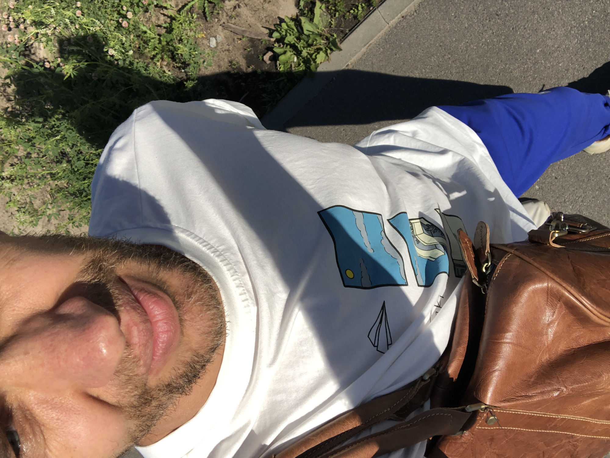 A man is taking a selfie outdoors in Saint Petersburg, Russia. He is wearing a white t-shirt with a cartoon graphic and bright blue sweatpants. A brown leather bag is slung over his shoulder. The background shows a sidewalk, vegetation, and shadows cast by sunlight. The man has short brown hair and a beard. He is looking down at the camera with a slight smile.