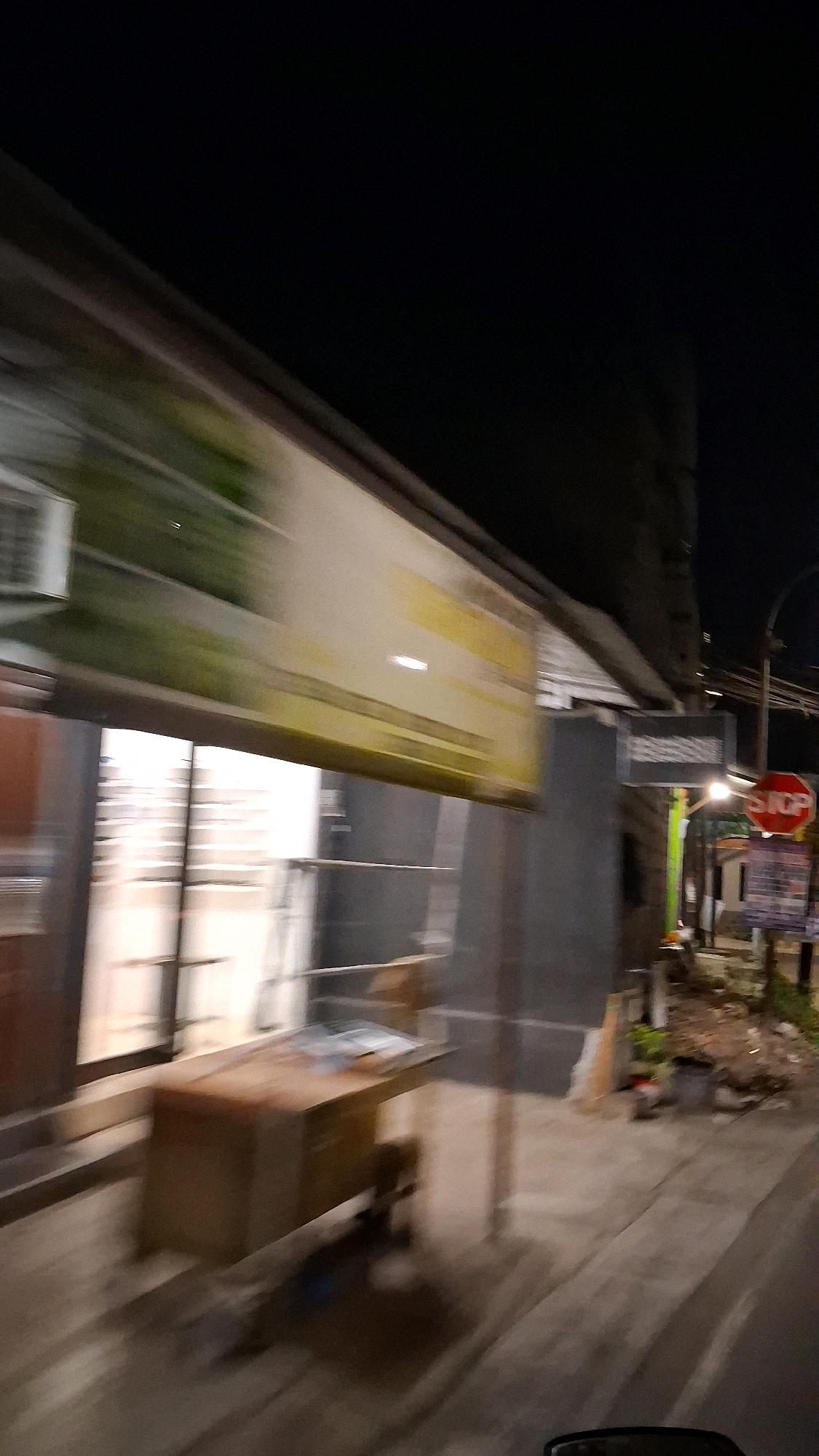 This image, captured at night in Berbah, Indonesia, shows a motion-blurred street scene. In the foreground, a brown wooden desk with what appears to be papers on it is visible through a glass storefront, suggesting the interior of a shop or office. To the right of the storefront, a red octagonal stop sign is prominently displayed on a pole, indicating a traffic intersection. Beyond the stop sign, there are other signs and some debris, typical of a roadside environment in a city. The overall lighting suggests artificial streetlights or lights from the businesses, creating a somewhat hazy atmosphere due to the motion blur. The dark sky above indicates it is late at night.