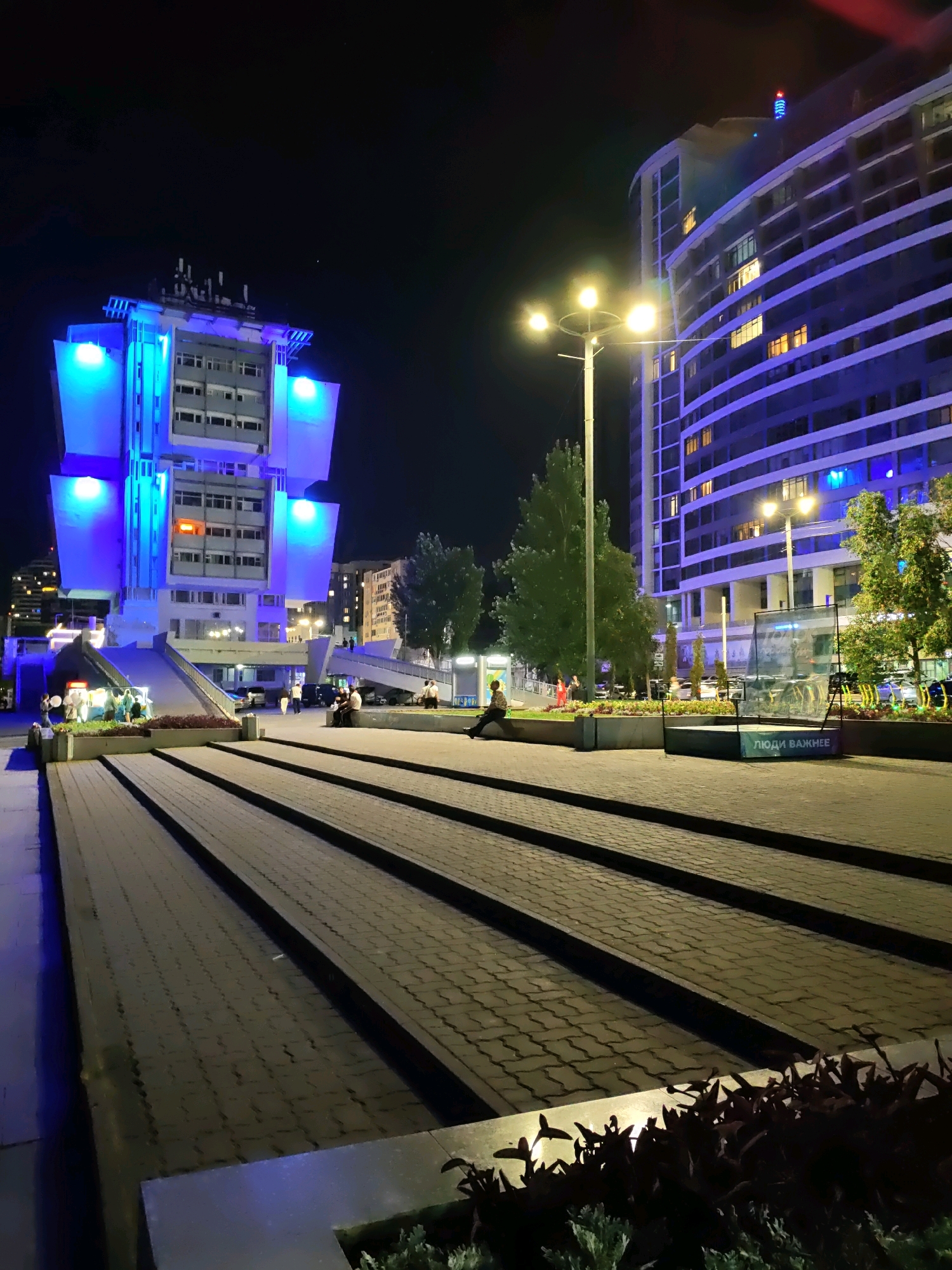 The image depicts a night scene in the city of Rostov-on-Don, Russia, specifically in front of the Central Market (Centralny Rynok) on Budennovsky Prospekt. In the foreground, a wide paved walkway with distinct lines is visible, leading towards a multi-level building illuminated with bright blue lights. This building appears to be a modern, somewhat angular structure. To the right, a taller, curved building with numerous windows, some lit, is also illuminated with blue light. Streetlights with multiple lamps are present, casting a warm glow. Several trees with lush green foliage are interspersed throughout the scene.
In the mid-ground, people are visible walking and sitting. One person is seated on the edge of the paved area on the right side of the frame, facing away from the camera. Further back, near the illuminated building, a group of people are gathered, some walking, and others appearing to be interacting. A glass enclosure with some objects inside and text that reads "ЛЮДИ ВАЖНЕЕ" (People are more important) is also visible. The ground is paved with rectangular bricks, and the overall lighting suggests a commercial or public urban space at night.