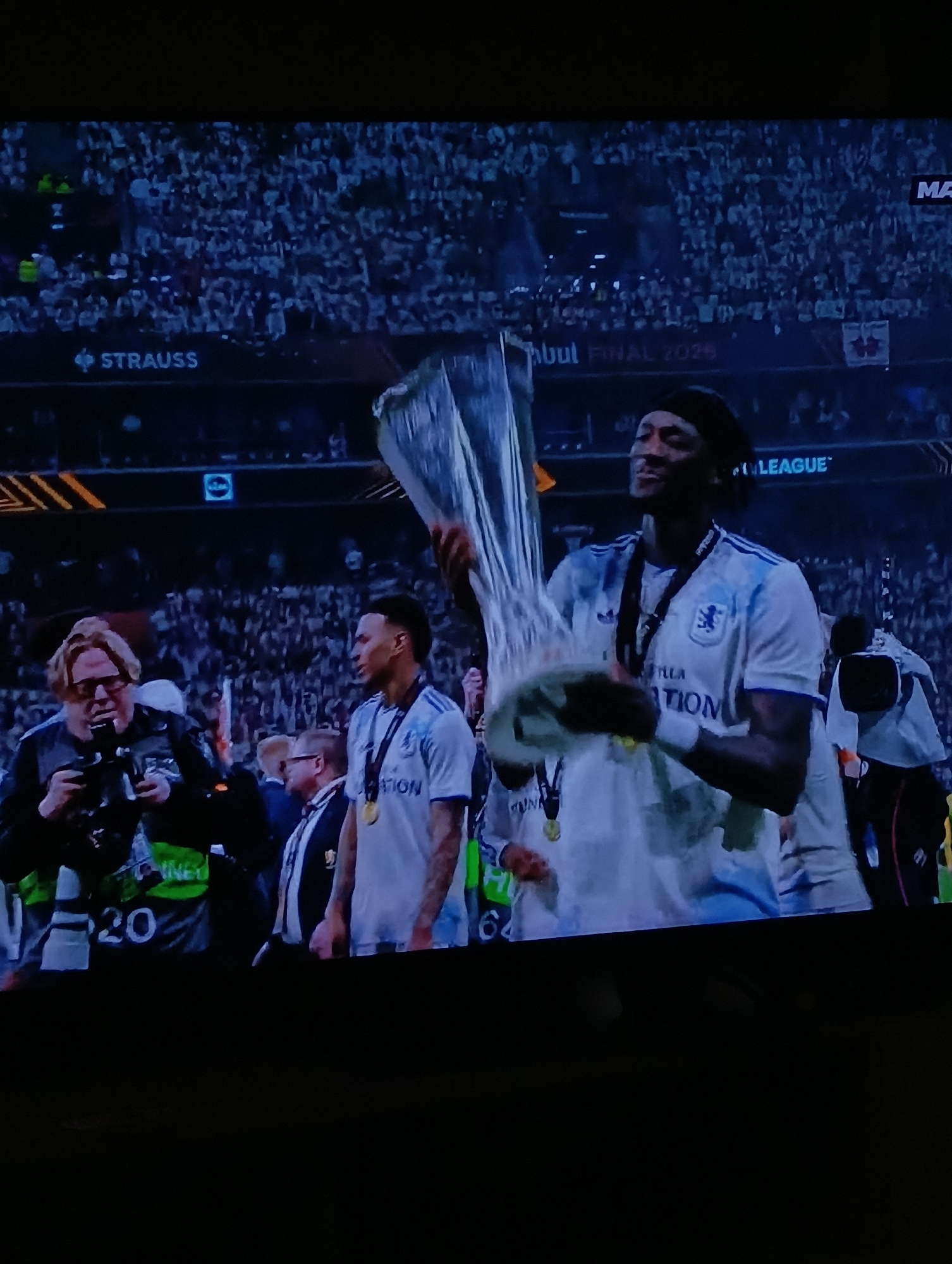 The image shows a man holding up a trophy. The setting appears to be after a sporting event, possibly a football (soccer) match, given the trophy and crowd in the background. The man holding the trophy is wearing a white jersey with the word "TION" visible on the front. He also has a medal around his neck. Several people are standing around him, including photographers. There is signage in the background, "Istanbul FINAL 2023", suggesting the event is related to the UEFA league. The location appears to be a stadium in Istanbul, Turkey. There is no evidence to suggest this occurred in Plovdiv, Bulgaria.