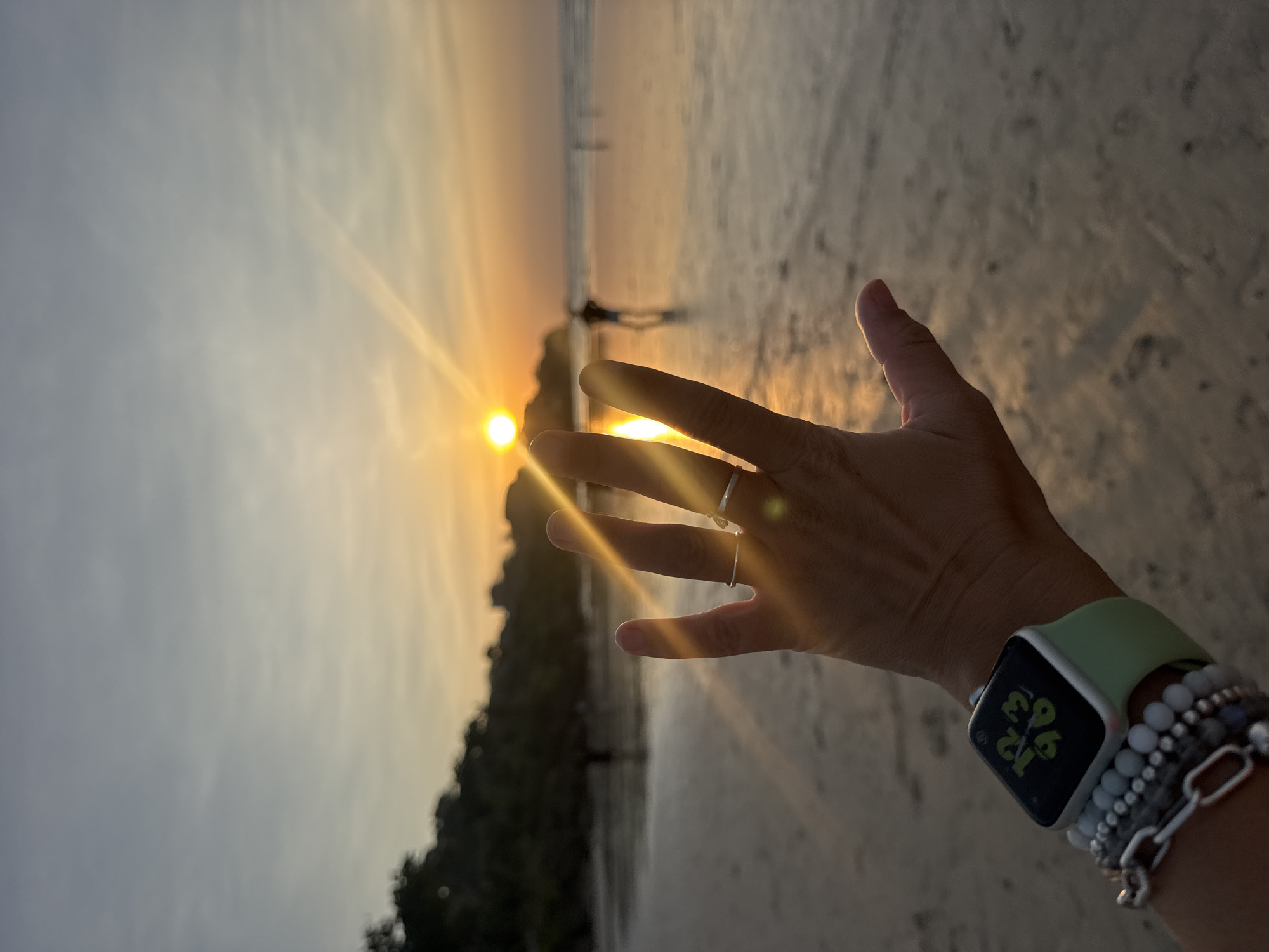 In the city of Kuta Selatan, Indonesia, at sunset on a beach, a hand with rings and bracelets is held up, blocking the view of the sun, which is low on the horizon and casting golden light across the water and wet sand. The sky is a gradient of pale blue and orange. Trees line the background. Several people are visible as silhouettes in the distance on the beach and in the water. The hand wears a smartwatch with a green band and a digital display showing time and other data.