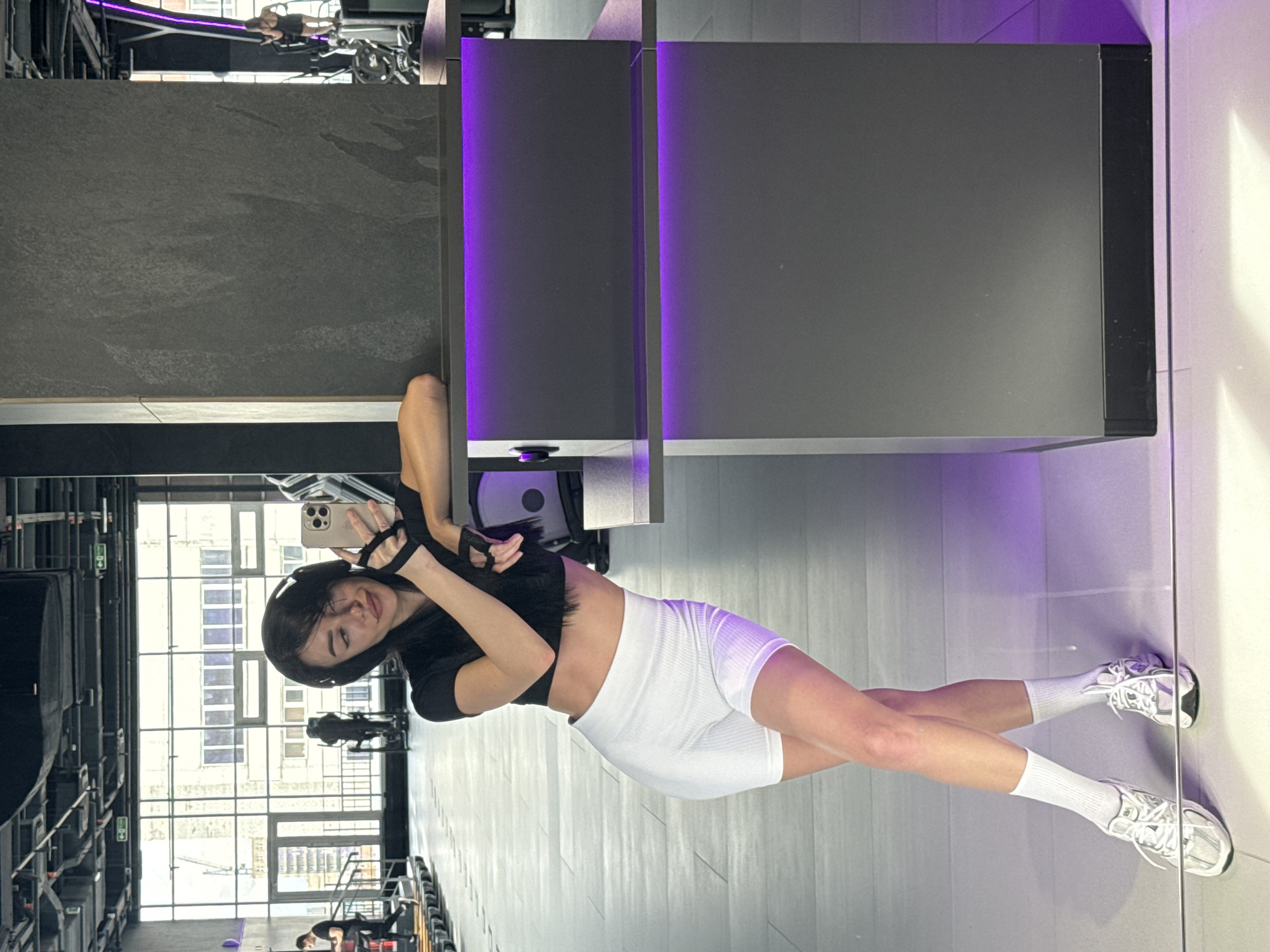 A woman is taking a selfie in a gym. She is wearing a black top, white shorts, and white socks and sneakers. She is holding her phone in her left hand. The gym is located in Krasnoyarsk, Russia. The gym has large windows with a view of a city building. There are exercise machines in the background.