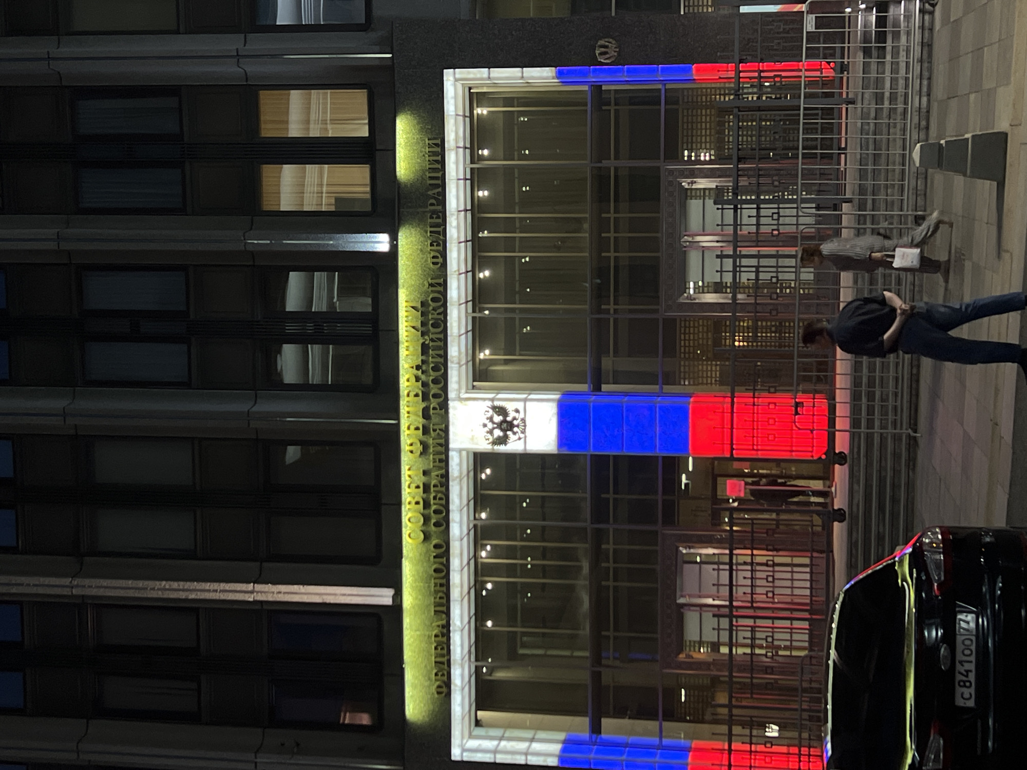 Here's a description of the image:
The image depicts the Federation Council building, located in Moscow, Russia, at night. The building's facade is partially illuminated, featuring the Russian tricolor flag (white, blue, red) along the upper and lower edges of the building's entrance. The inscription "СОВЕТ ФЕДЕРАЛЬНОГО СОБРАНИЯ РОССИЙСКОЙ ФЕДЕРАЦИИ" (Council of the Federal Assembly of the Russian Federation) is visible in green on the building's exterior.
Two pedestrians are near the entrance. A black car is parked along the edge of the frame, partially obscuring the view of the building. The car's license plate number, C841OO77, is partially visible. A metal barricade is in place in front of the building. The rest of the building appears to be a standard multi-story building.