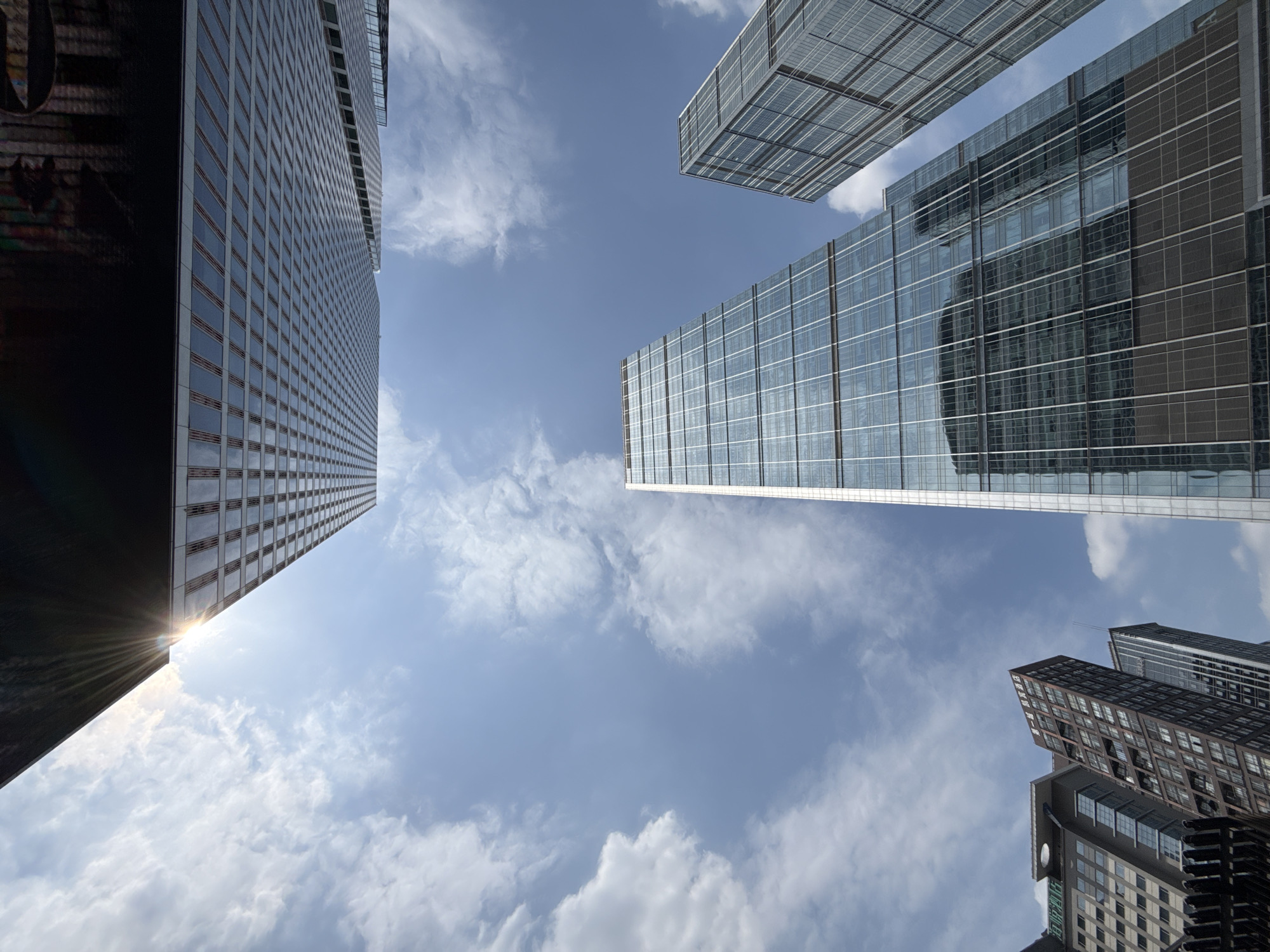 The image shows a low-angle view of several tall buildings under a cloudy blue sky. The buildings have a modern architectural style, with glass facades and grid-like patterns. One building on the left reflects sunlight. The presence of these modern buildings suggests an urban setting, consistent with the Jinjiang District in China. There are no people visible in the image. The focus is on the architecture and the sky.