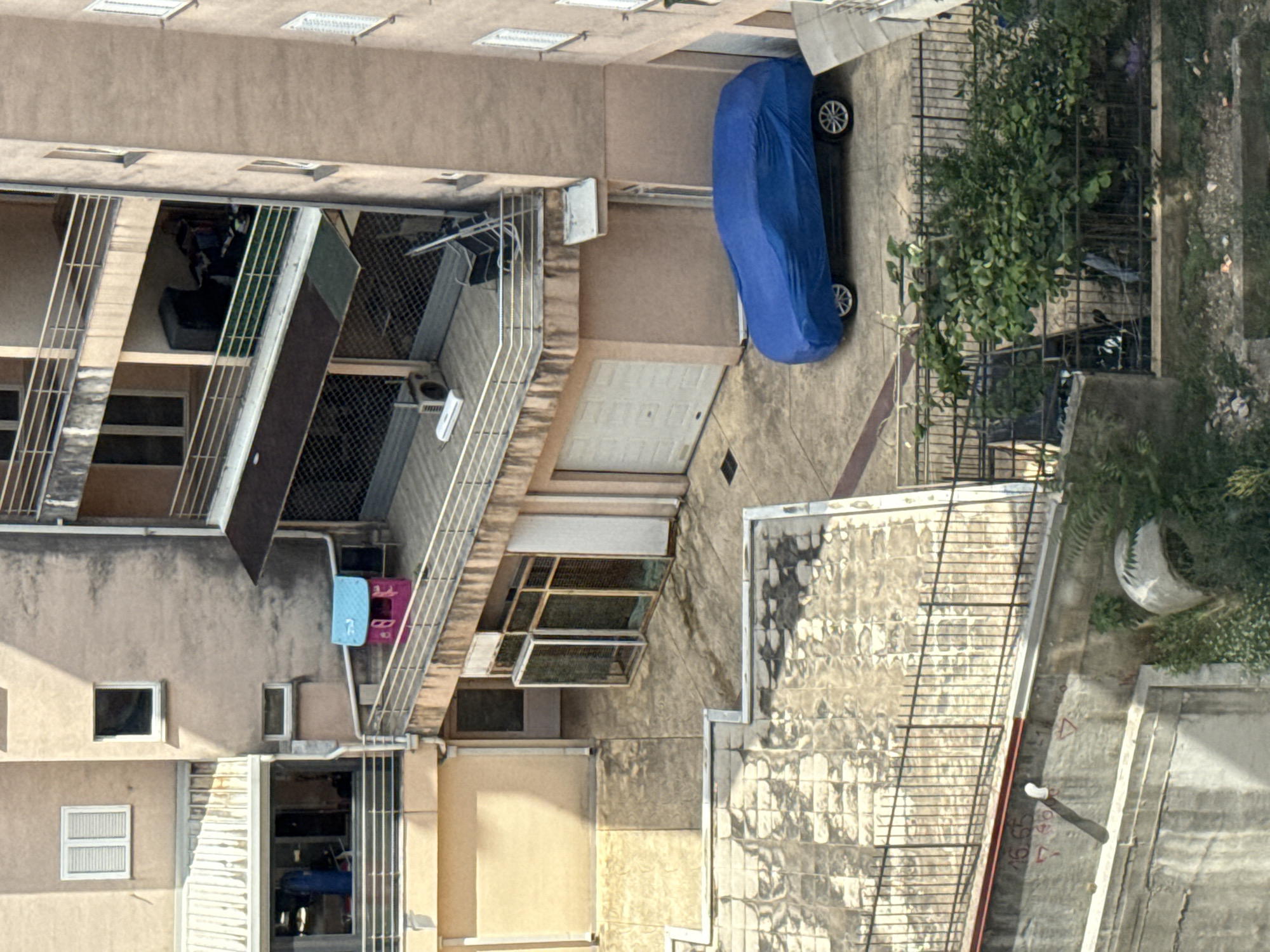 The image depicts an aerial view of an apartment building in Budva, Montenegro. In the foreground, a blue car cover encloses a vehicle parked on a concrete surface. The building features multiple balconies with railings, one of which has a small pink and blue playhouse. A closed white garage door is visible at the ground level. Further down, a section of a fenced terrace with overgrown greenery and what appears to be a discarded concrete structure is present. Markings, possibly related to construction or utilities, are visible on a lower concrete wall. The overall scene suggests a residential area with a mix of urban elements and outdoor spaces.