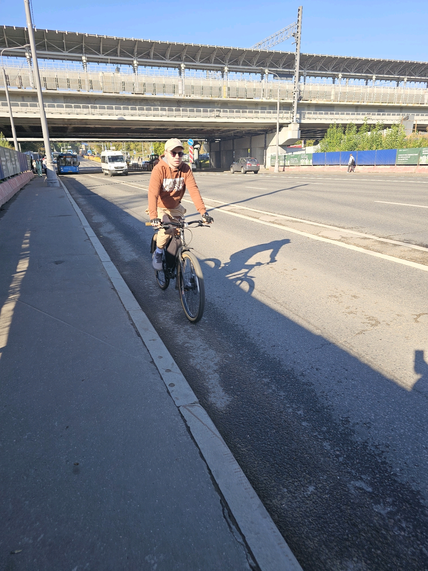 The image captures a person riding a bicycle on a sunny day in Moscow, Russia. The cyclist, wearing a light brown hooded sweatshirt and a beige baseball cap, is moving along a paved pedestrian path adjacent to a multi-lane road. The road has several vehicles, including a bus and a white van, visible in the background, indicating urban traffic. Overhead, a large elevated structure, likely a railway or highway overpass, dominates the upper portion of the scene. The sky is clear and blue. The sunlight casts a long shadow of the cyclist and their bicycle onto the road. The environment appears to be a typical urban setting with infrastructure and transportation elements characteristic of Moscow.