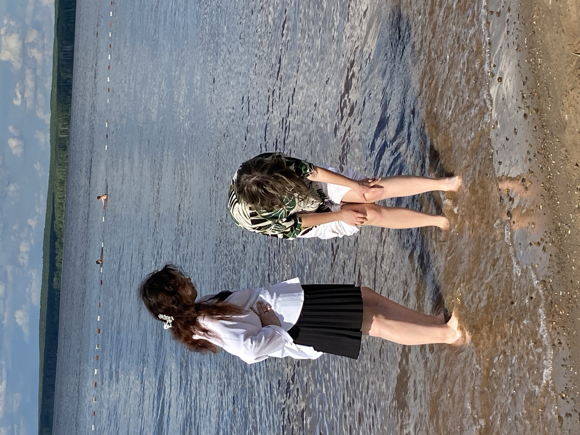 Two individuals are standing at the edge of a body of water, which is likely a reservoir or a large river, in Chaikovsky, Russia. The water is calm with gentle waves lapping the shore. A sandy beach with scattered pebbles is visible in the foreground. In the background, a line of buoys separates the swimming area from the deeper water, and a forested shoreline can be seen in the distance under a cloudy sky.
The individual on the left, facing the water, is wearing a white open-collared shirt over a dark top and a black pleated skirt. Their hair is dark and curly, adorned with a white flower clip. Their left hand is resting on their hip.
The individual on the right is bending over, with their hands on their knees, facing the water. They are wearing a patterned short-sleeved shirt in green and white and white shorts. Their hair is medium length and appears to be light brown.
Two other figures are visible in the water in the background, suggesting recreational swimming. The scene depicts a casual outdoor setting on a warm day.