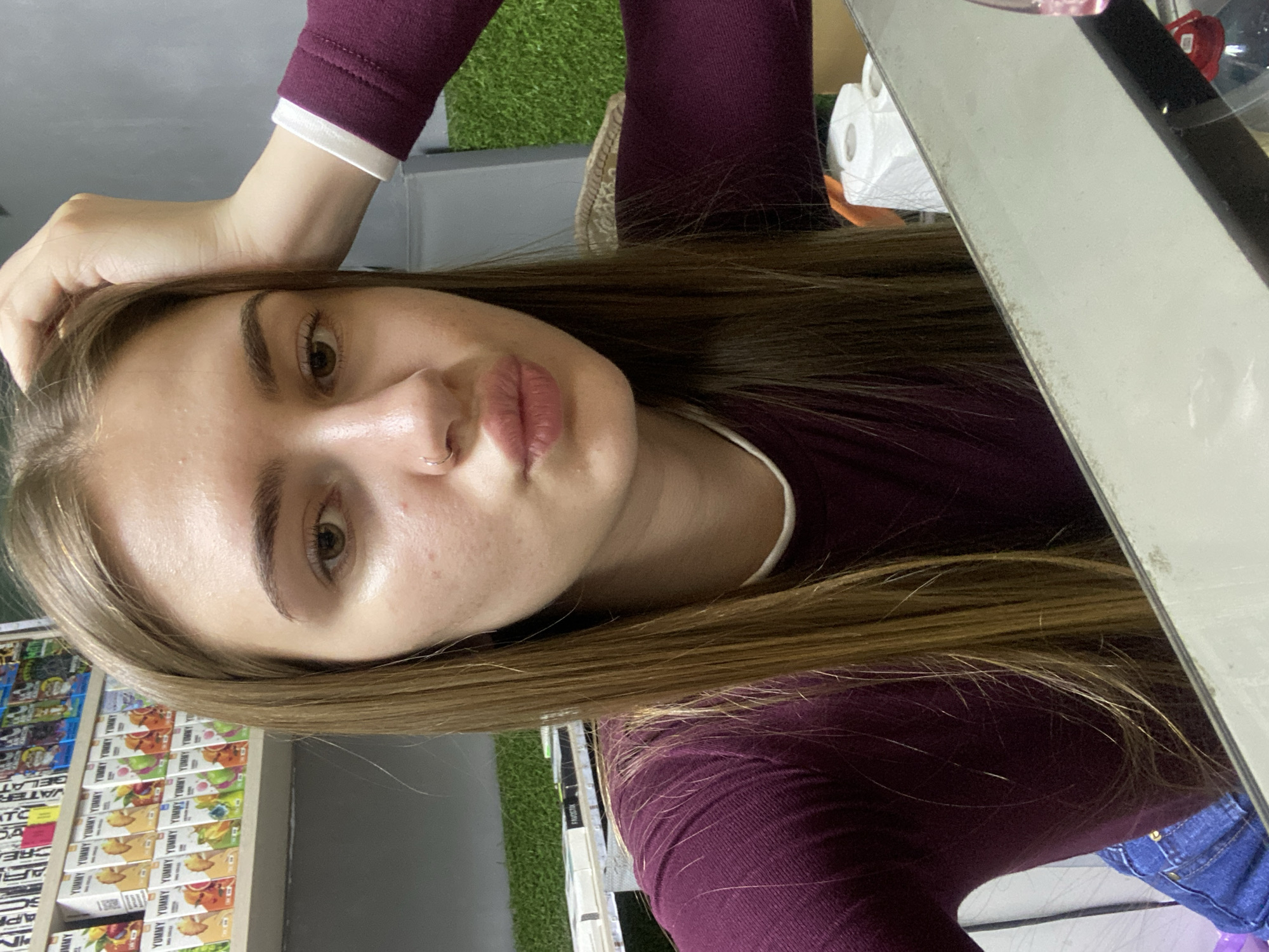 A young woman with long, brown hair and a nose ring is taking a selfie in what appears to be a retail establishment. She is wearing a maroon long-sleeved shirt and blue jeans. She has her right hand on her head. Behind her, shelves are stocked with various products, some of which are colorful boxes with graphics. To her right, there is a section of artificial grass. In the foreground, a reflective surface, possibly a counter or mirror, is visible. The environment suggests a store or kiosk, possibly selling vape products or similar small goods, located in the city of Dyakovo, Russia.