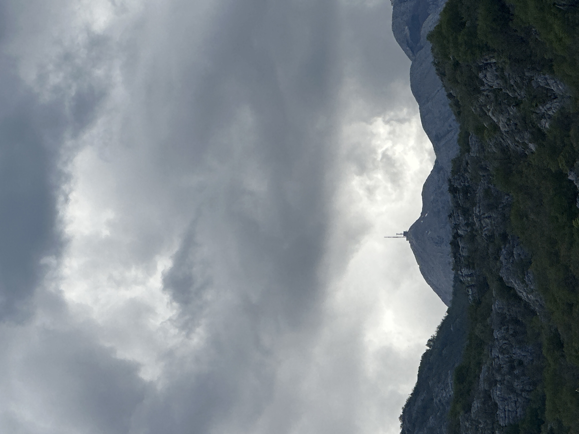 The image shows a mountain peak in Očinići, Montenegro, partially obscured by clouds. A transmission tower is visible at the summit. The mountain's slope is covered in vegetation. The sky is overcast with grey clouds.
