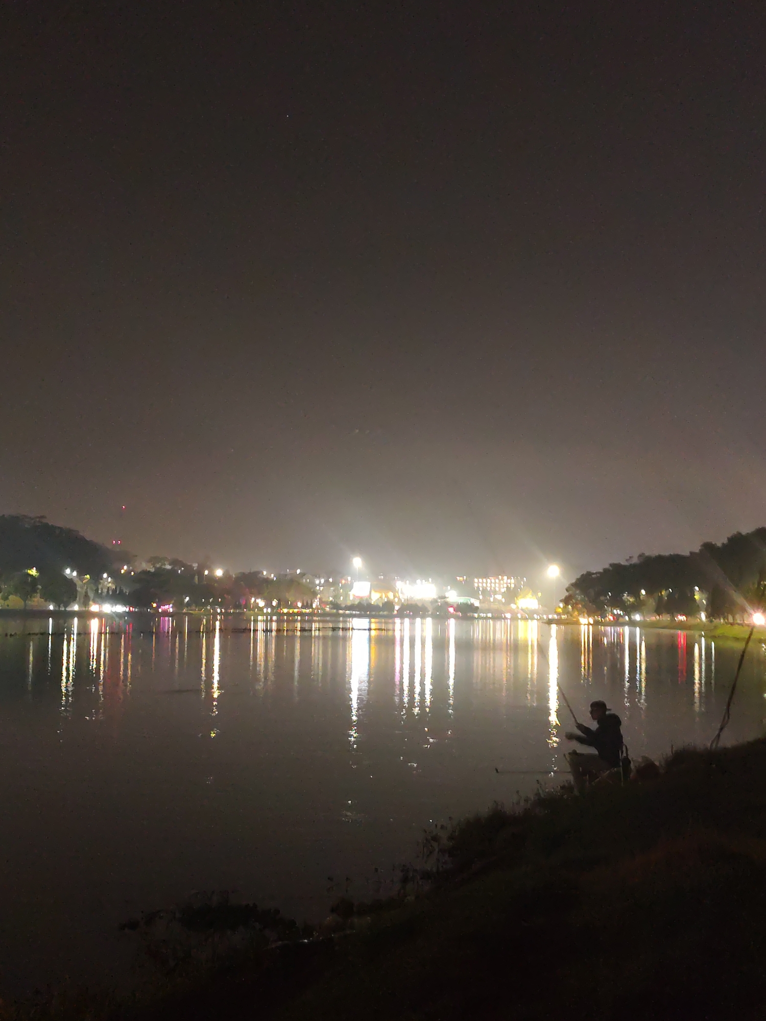 At night, a person is fishing in Ho Xuan Huong Lake in Da Lat, Vietnam. The lake reflects the lights of the city. The person is standing on the edge of the lake, holding a fishing rod. The city lights in the background are blurred and indistinct.