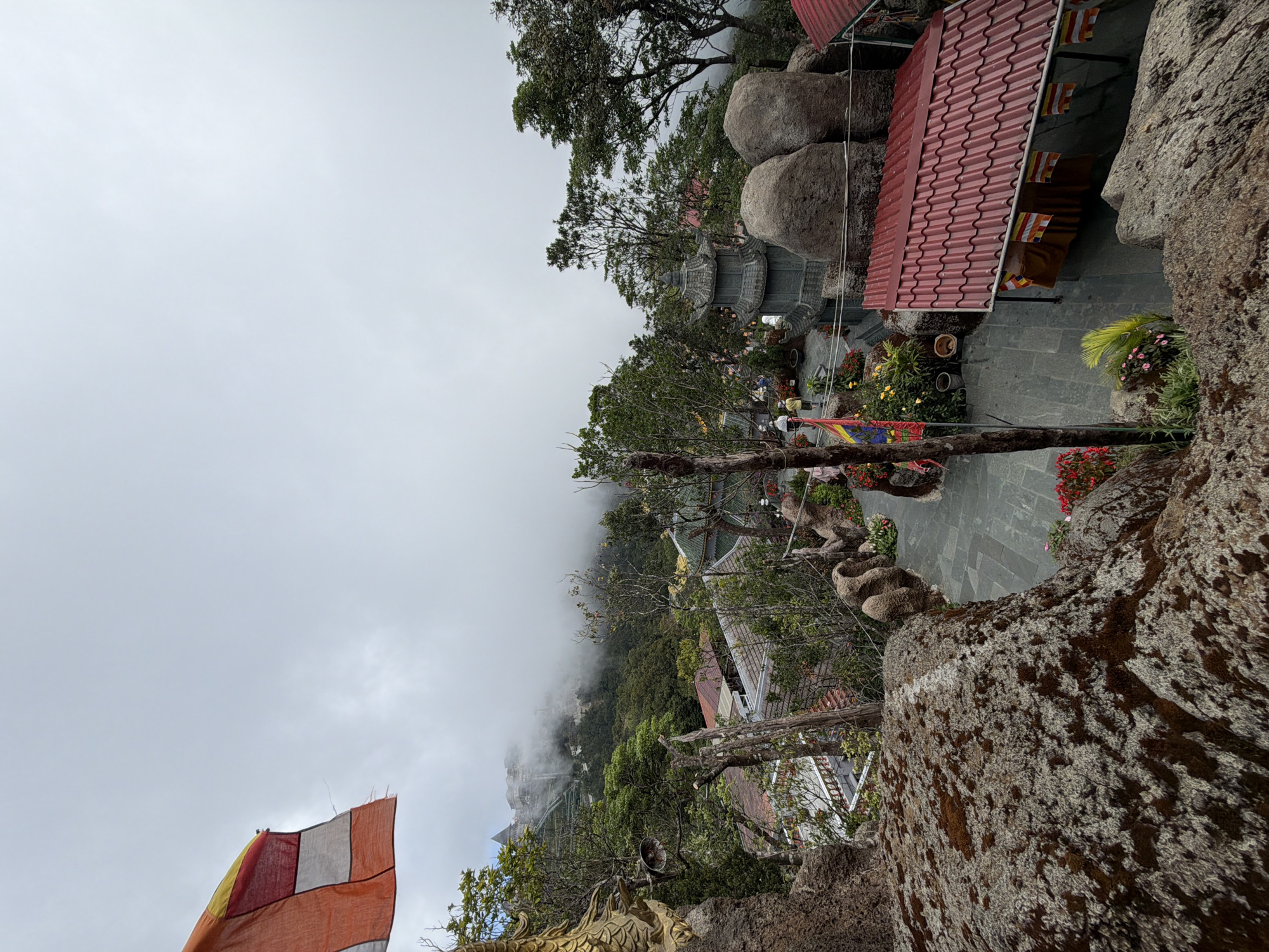 This is a high-angle, full shot of a misty mountain landscape with traditional Vietnamese architecture. In the foreground, large moss-covered rocks dominate the left side of the frame, with a golden dragon statue visible on the far left. A flag with multiple colored panels flutters from a pole.
Further into the scene, a paved pathway winds through a garden area with potted plants and colorful flowers. A multi-tiered pagoda stands to the right of the path, flanked by large boulders. Beyond this, a red-roofed building with an overhang is visible. In the background, the landscape descends into a valley shrouded in thick white mist, with glimpses of trees and the distant outlines of buildings, suggesting a mountainous resort or temple complex. Two figures in casual clothing are walking on the path.
The scene is likely located at the Bà Nà Hills resort area in Đà Nẵng, Vietnam, known for its French colonial village, gardens, and spiritual sites atop a mountain. The overcast and misty conditions are typical for this elevated location.