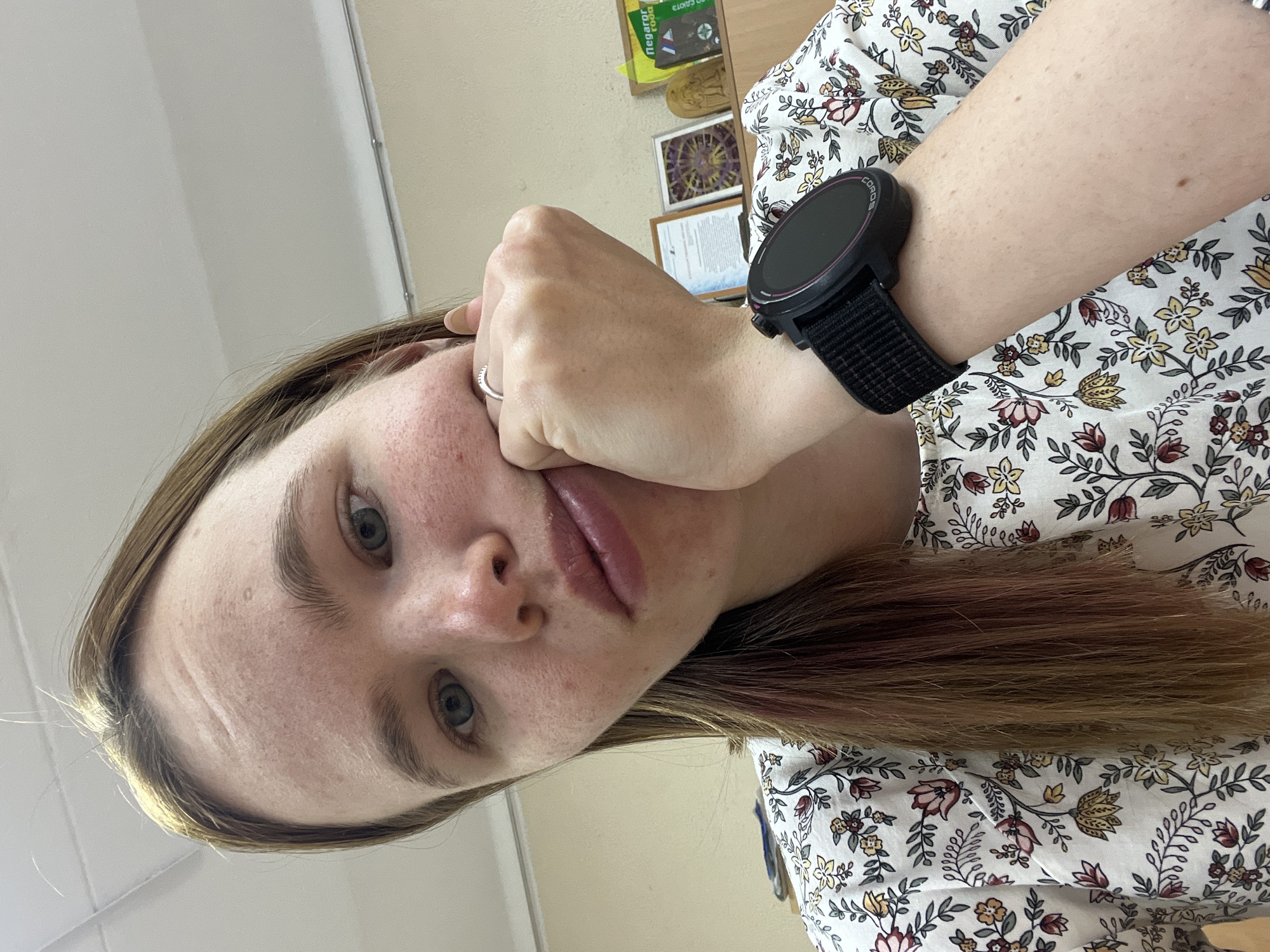 A young, fair-skinned woman is shown in a close-up, angled selfie. Her hand is pressed against her cheek. She has light brown hair, and wears a floral print shirt. A black smartwatch is visible on her wrist. The background appears to be an indoor setting with a light-colored wall and a shelf or desk with various items including framed images. The location is Chaikovsky, Russia.