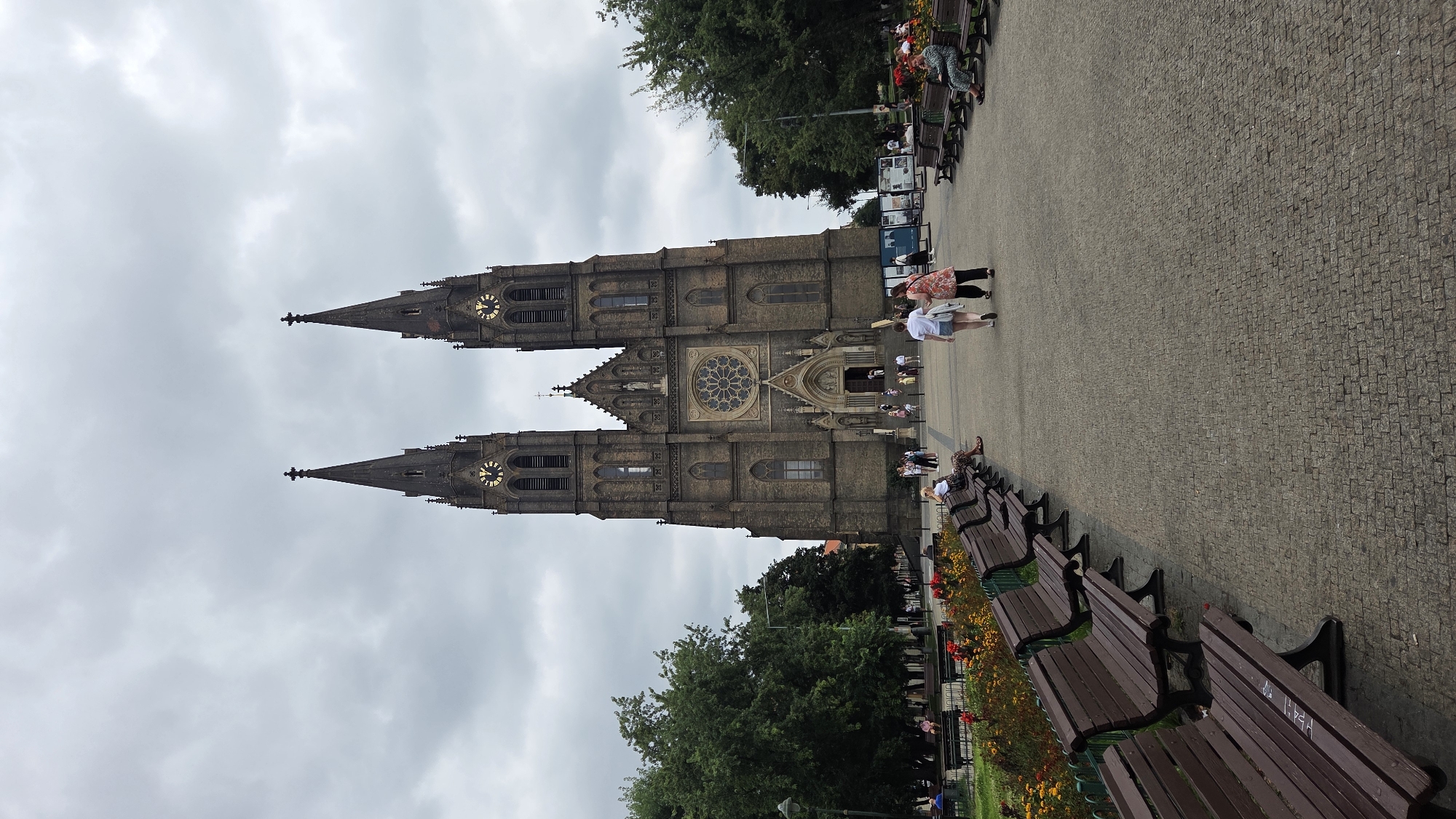 The image shows the Basilica of St. Peter and St. Paul in Vyšehrad, Prague, Czechia, on an overcast day. The imposing brick structure features two tall spires, each topped with a cross, and large clock faces. In front of the basilica, a paved plaza stretches forward, lined with rows of dark wooden benches on either side. To the left of the plaza, a garden area with vibrant red and yellow flowers is visible, separated by a green fence. Several people are present in the scene: some are walking along the plaza, including two women in the foreground; others are seated on the benches, observing the surroundings or resting. Trees and greenery frame the sides of the plaza, suggesting a park-like setting.