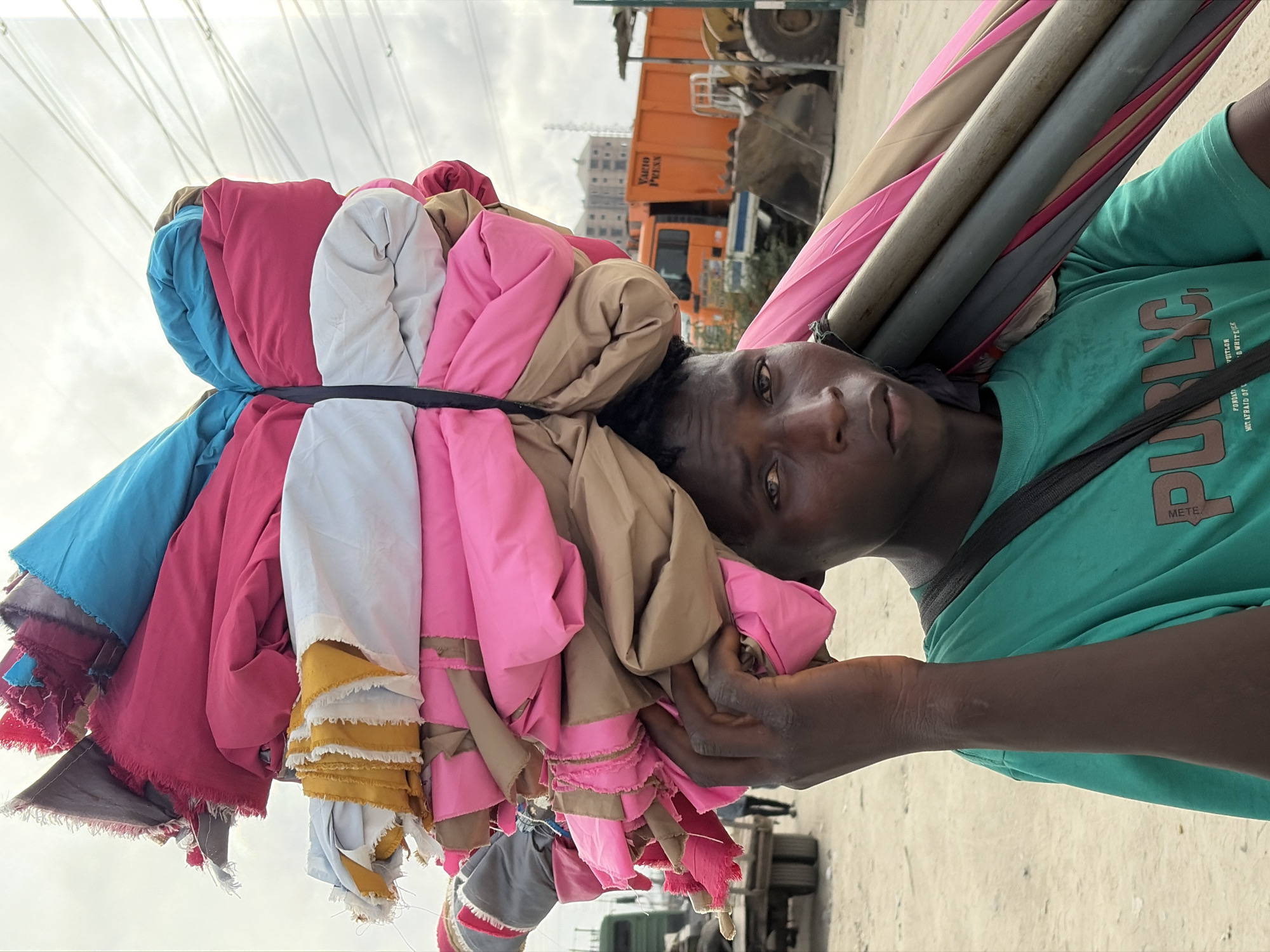 A young Black male in Igboefon, Nigeria, carries a large bundle of rolled fabrics and what appear to be umbrella poles on his head and shoulder. He is wearing a green t-shirt with the word "PUBLIC" printed on it in brown lettering. The fabrics are of various colors, including pink, blue, white, and tan. In the background, there are utility poles and lines, construction vehicles, and buildings. The ground is dirt and gravel, suggesting a possibly industrial or construction area. The overall scene depicts a vendor in an urban environment.