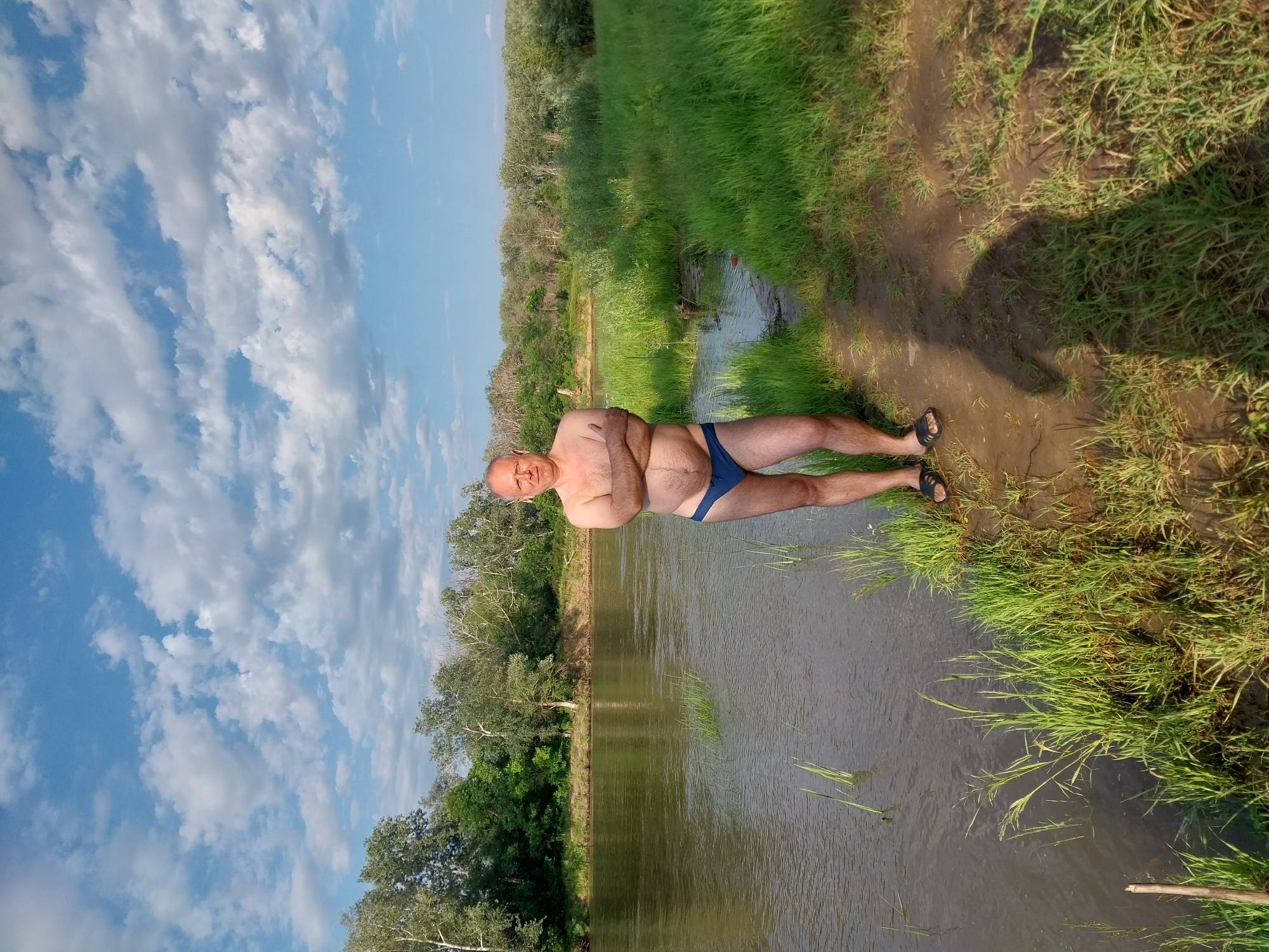 An adult male stands on the bank of a river, wearing blue swim briefs and black sandals. He has his arms crossed and is facing the camera with a neutral expression. The river is calm and reflects the blue sky with scattered white clouds. Lush green vegetation lines the opposite bank, with trees forming a dense backdrop. The foreground consists of a muddy bank with grass. The image appears to be taken outdoors during the daytime, likely in a rural or natural setting. The location is identified as Chkalov, Russia.