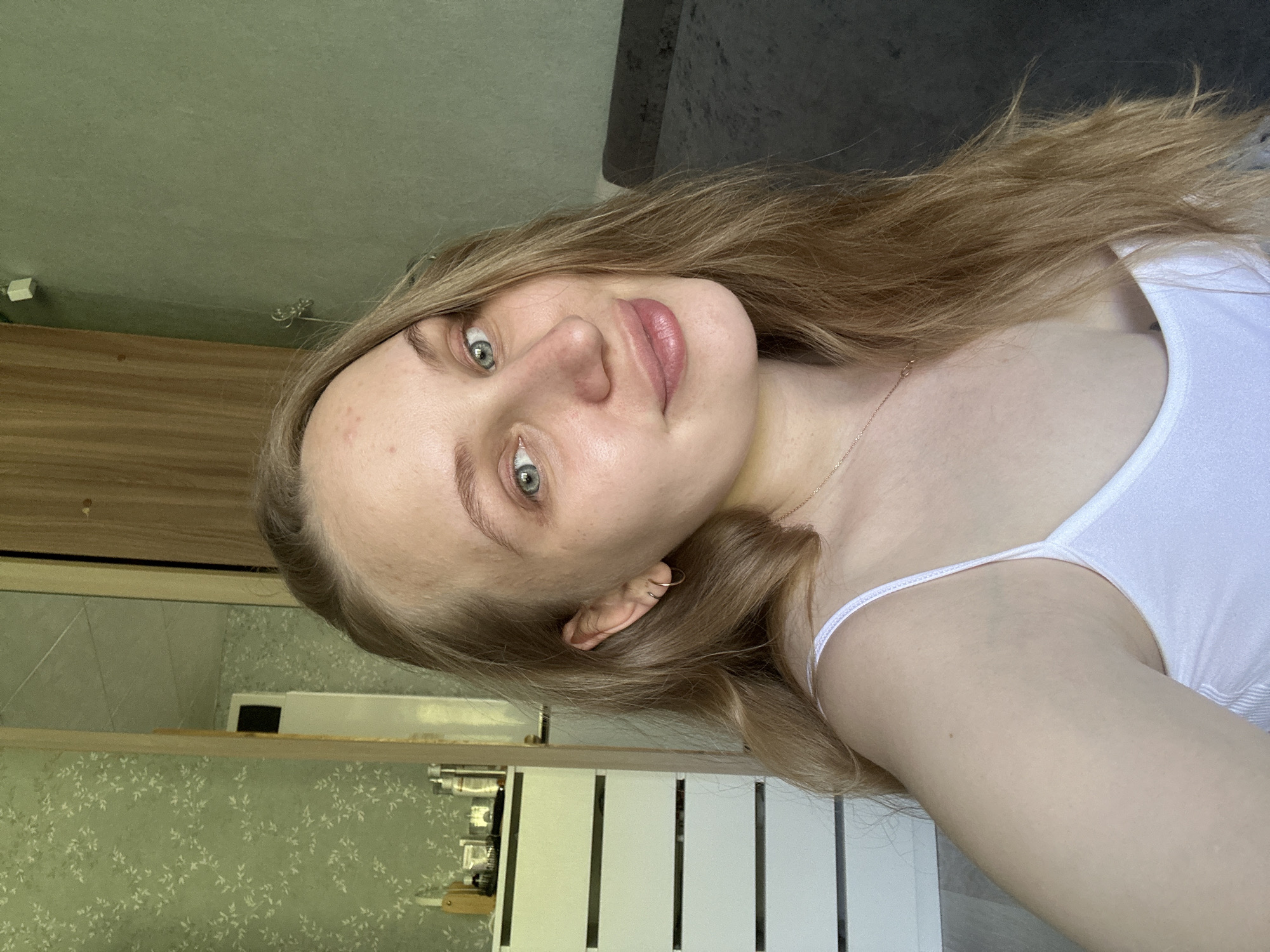 A fair-skinned woman with long, light brown hair and blue eyes is taking a selfie indoors. She is wearing a white tank top. The background includes a cabinet with white doors and a wallpapered wall with a light green and white floral pattern. It's impossible to determine the specific location within Chaikovsky, Russia, based solely on the image.