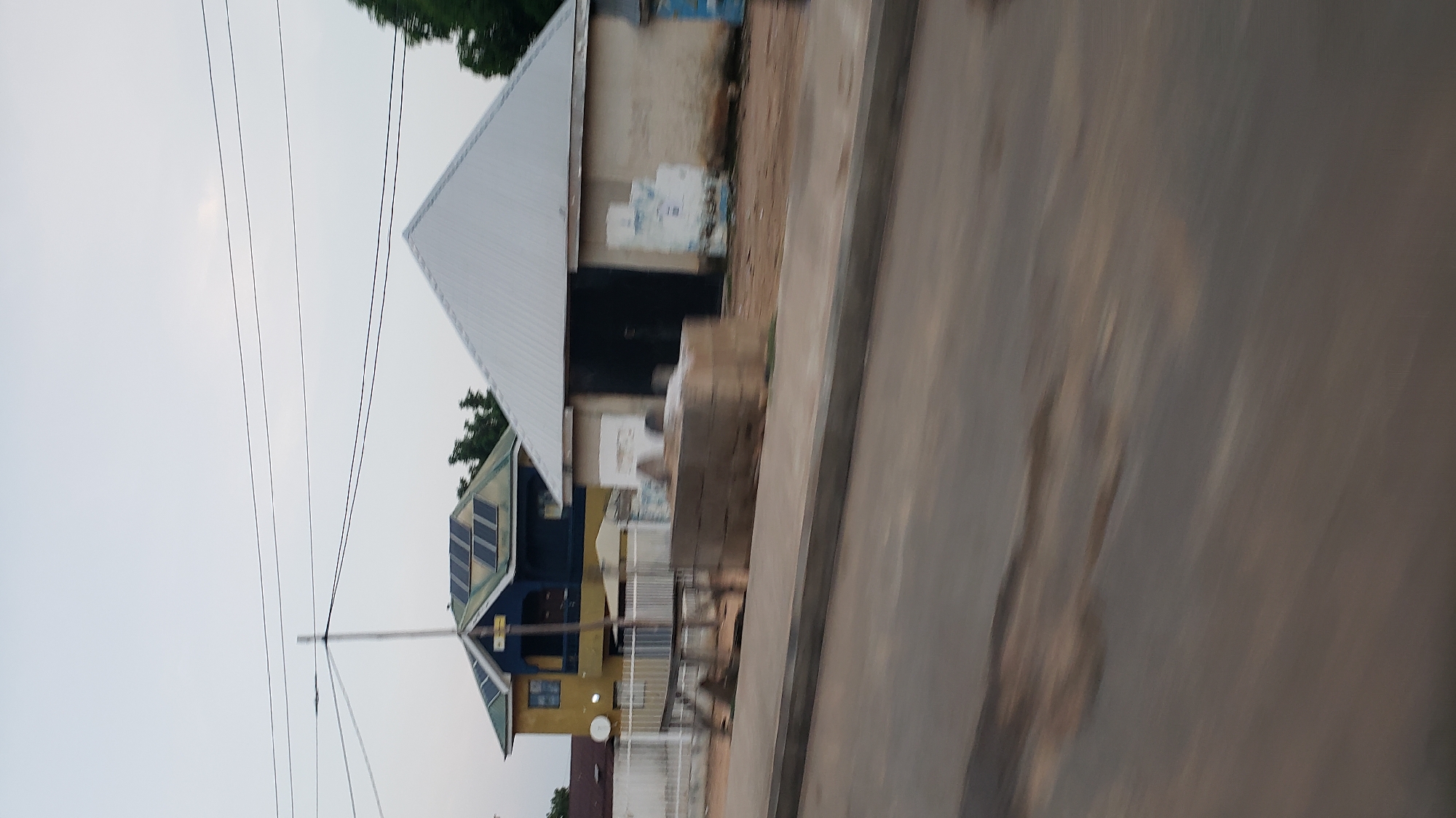 In Jalingo, Nigeria, a street scene unfolds with two distinct structures in the background. To the left, a two-story building, painted yellow and dark blue, features a gabled roof covered in solar panels. A balcony is visible on the second floor. To the right, a single-story building with a white, corrugated metal gabled roof stands adjacent, with a large, dark doorway. The area in front of these buildings appears to be a construction site or storage area, with a pile of cement blocks and a wooden structure. The foreground shows a blurred asphalt road with a gray concrete curb, indicating the photo was taken from a moving vehicle. The sky is overcast.