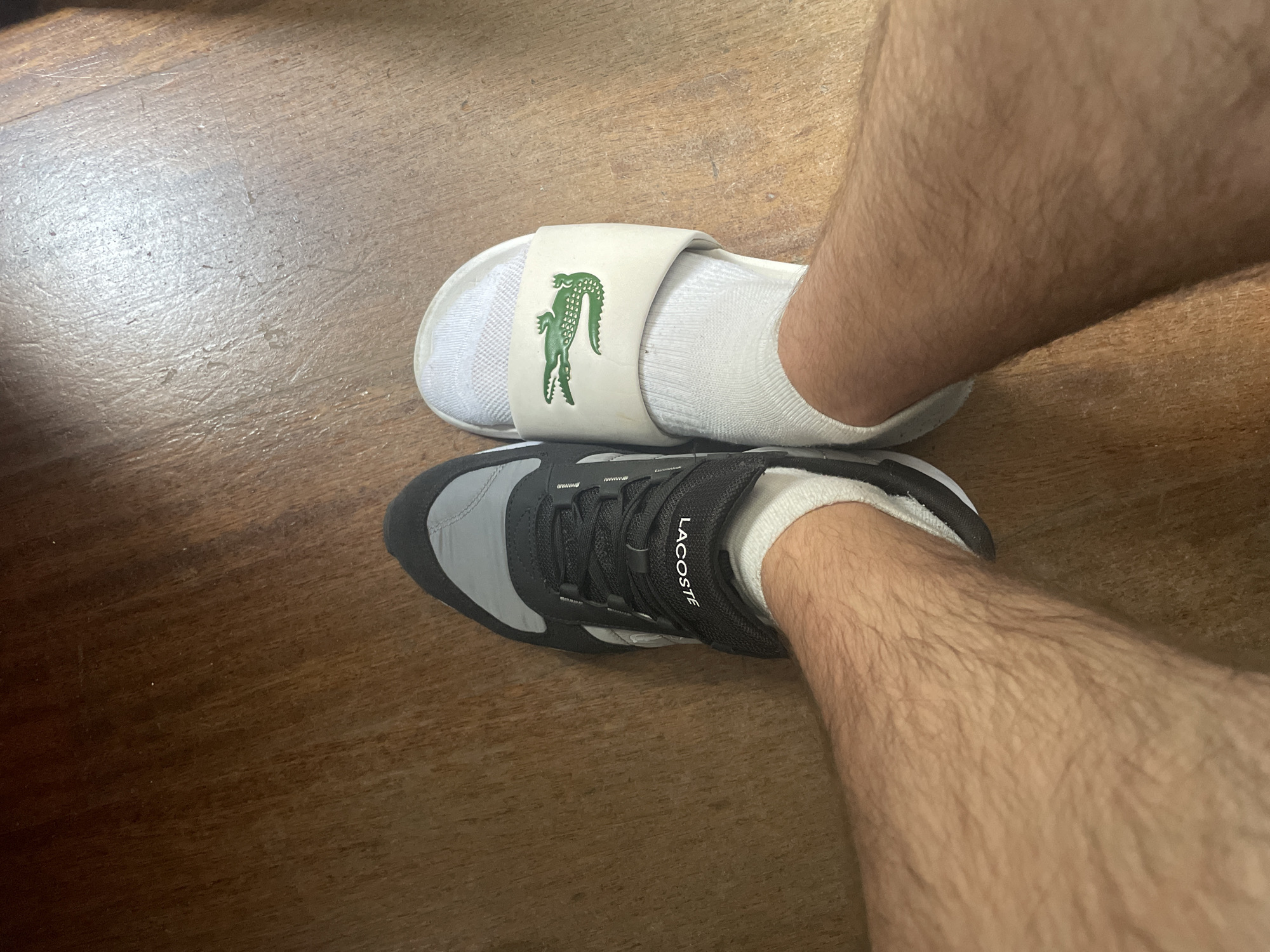The image shows a top-down view of a person's legs, with one foot wearing a black and gray Lacoste sneaker and the other foot wearing a white Lacoste slide sandal. Both feet are covered with white socks. The legs are hairy. The shoes are resting on a wooden floor with visible grain. The location is not specified in the image.