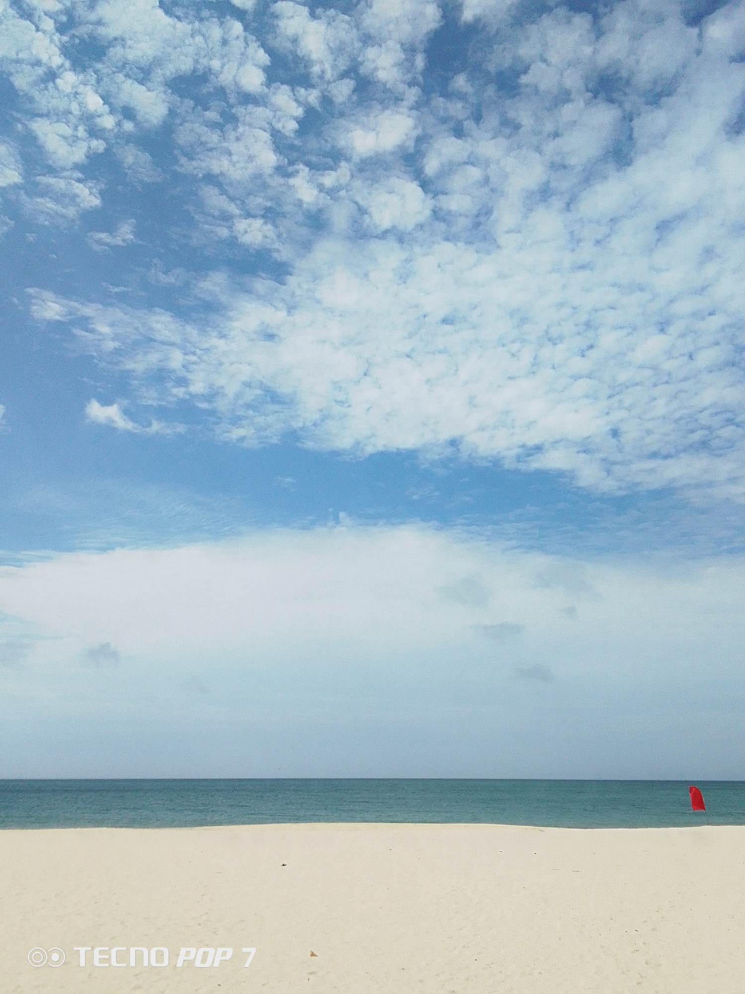 The image depicts a coastal scene in Pedro González, Venezuela. The foreground shows a wide sandy beach. Beyond the beach is the ocean, and above is a sky filled with white clouds. A single red flag is planted on the beach near the water's edge. There are no people visible in the scene. The horizon line appears flat and even. The sky takes up the top portion of the frame with a blue and white sky and the white sandy beach in the bottom.