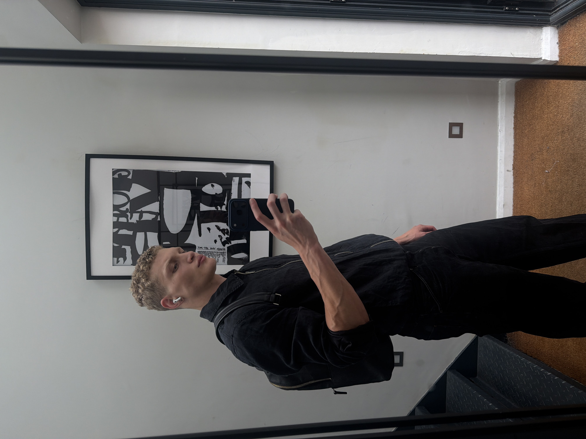 A young Caucasian male with short, curly blond hair is reflected in a full-length mirror, taking a selfie with his smartphone. He is wearing a black zip-up jacket over a black collared shirt, and black pants. He has a black backpack with leather straps slung over his left shoulder. He is also wearing white wireless earbuds. In the background, there is a black and white abstract art print framed on a light gray wall. To the left of the frame, a dark metal staircase with diamond plate treads is visible. The floor is covered with a brown, textured mat. The scene appears to be an indoor hallway or entryway. Based on the matte texture of the wall and the style of the light fixture on the wall, this could be a modern residential interior, potentially in a London neighborhood.